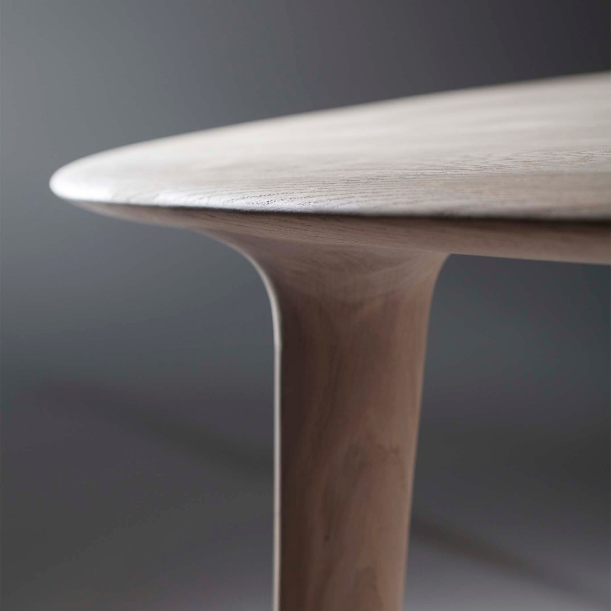 Close-up of the LUC Table by Artisan, showcasing its elegant, smooth design. The rounded leg seamlessly blends into the light wood top, highlighting its craftsmanship. A blurred gray background accentuates the exquisite finishes and tactile appeal.