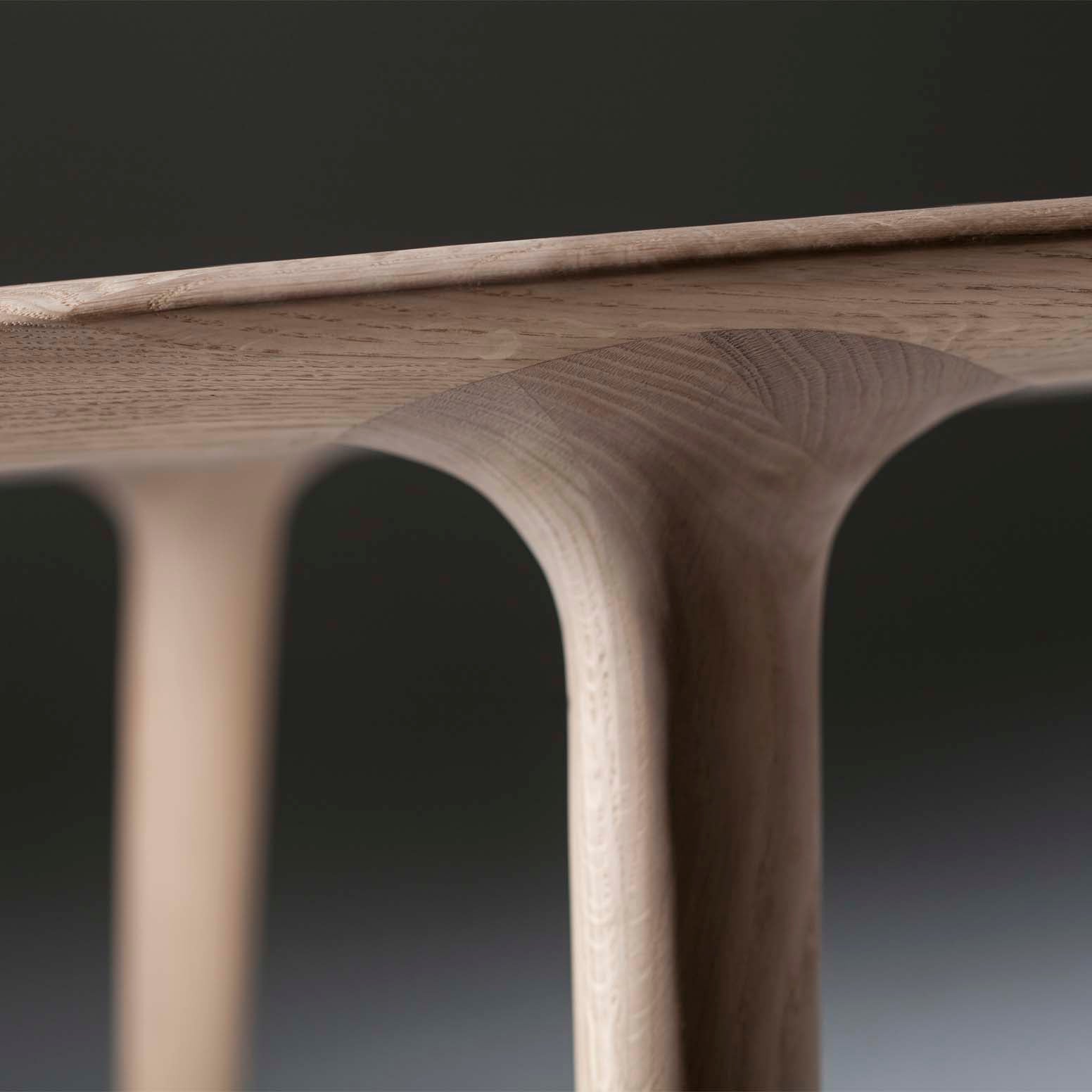 Close-up of the Artisan LUC Table highlighting its elegantly crafted wooden edge with smooth curves and natural wood grain. The artisan craftsmanship is evident in the tables arched structure, connecting the tabletop to its legs against a dark background.