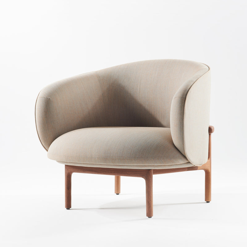 Part of the Artisan MELA Collection, the MELA Lounge Chair Trimmed features a modern, light gray upholstery with a curved backrest and soft padding. With wooden legs and a sleek, mid-century inspired design, it elegantly stands against a white background.