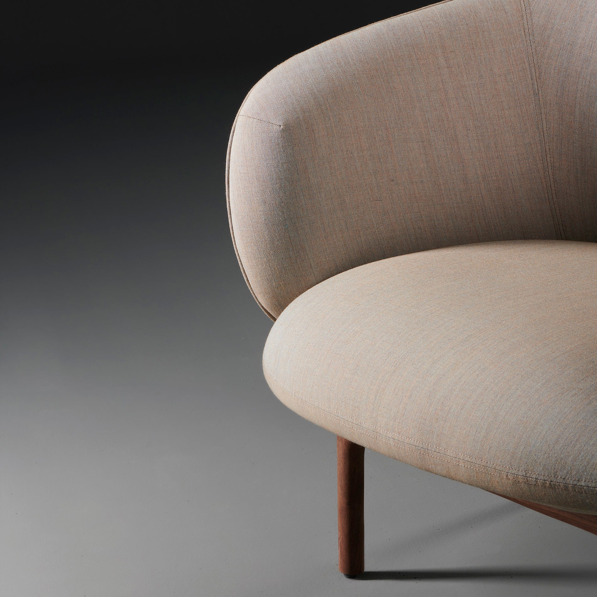 Close-up of the Artisan MELA Lounge Chair Trimmed, showcasing its modern beige upholstery and mid-century style curved backrest on wooden legs against a dark gray background, highlighting its elegant minimalist design that embodies comfort and style.