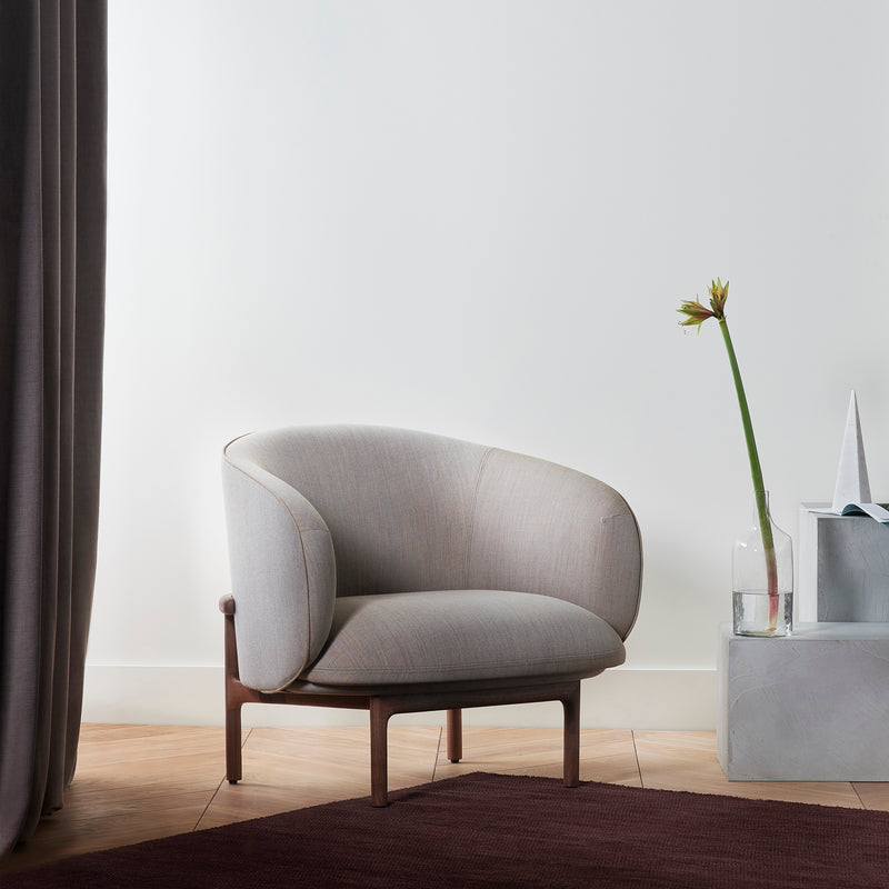 An Artisan MELA Lounge Chair Trimmed with a beige upholstered shell and wooden legs sits on a wooden floor near a light gray wall. Next to it is a minimalist concrete stand with a glass vase holding an orange-petaled flower. A dark rug partially covers the floor.