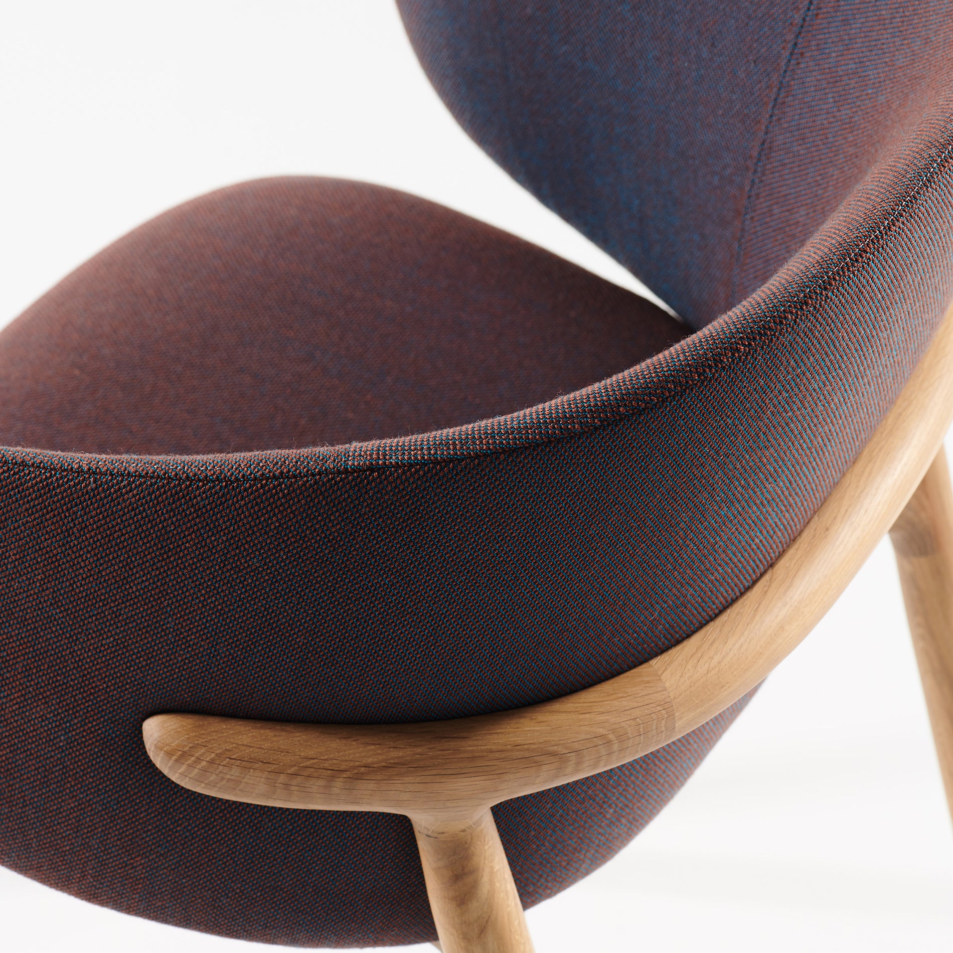 Close-up of the Artisan Mela Chair featuring textured dark fabric upholstery in blue and brown. It has a curved wooden frame made of light wood, highlighting its sleek mid-century minimalist design with an elegantly upholstered backrest.