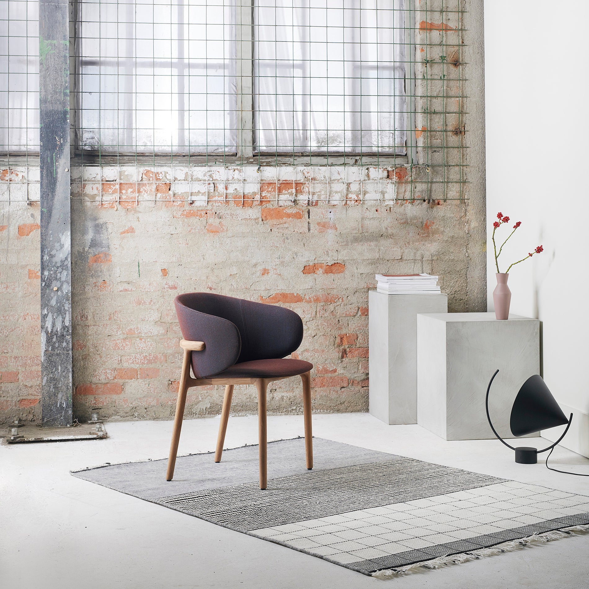 In a modern room, an Artisan Mela Chair with a purple upholstered backrest sits on a gray and white rug, against a brick wall with a large window. A concrete block displays stacked books and red flowers in a vase, while a unique black floor lamp exudes mid-century charm nearby.