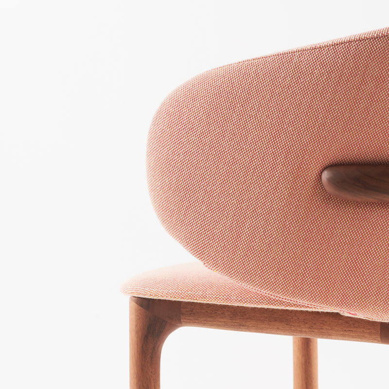 A close-up of the Artisan Mela Chair showcases its elegant contours with a curved, cushioned backrest in soft pink fabric and a sleek mid-century wooden frame and legs set against a white background.