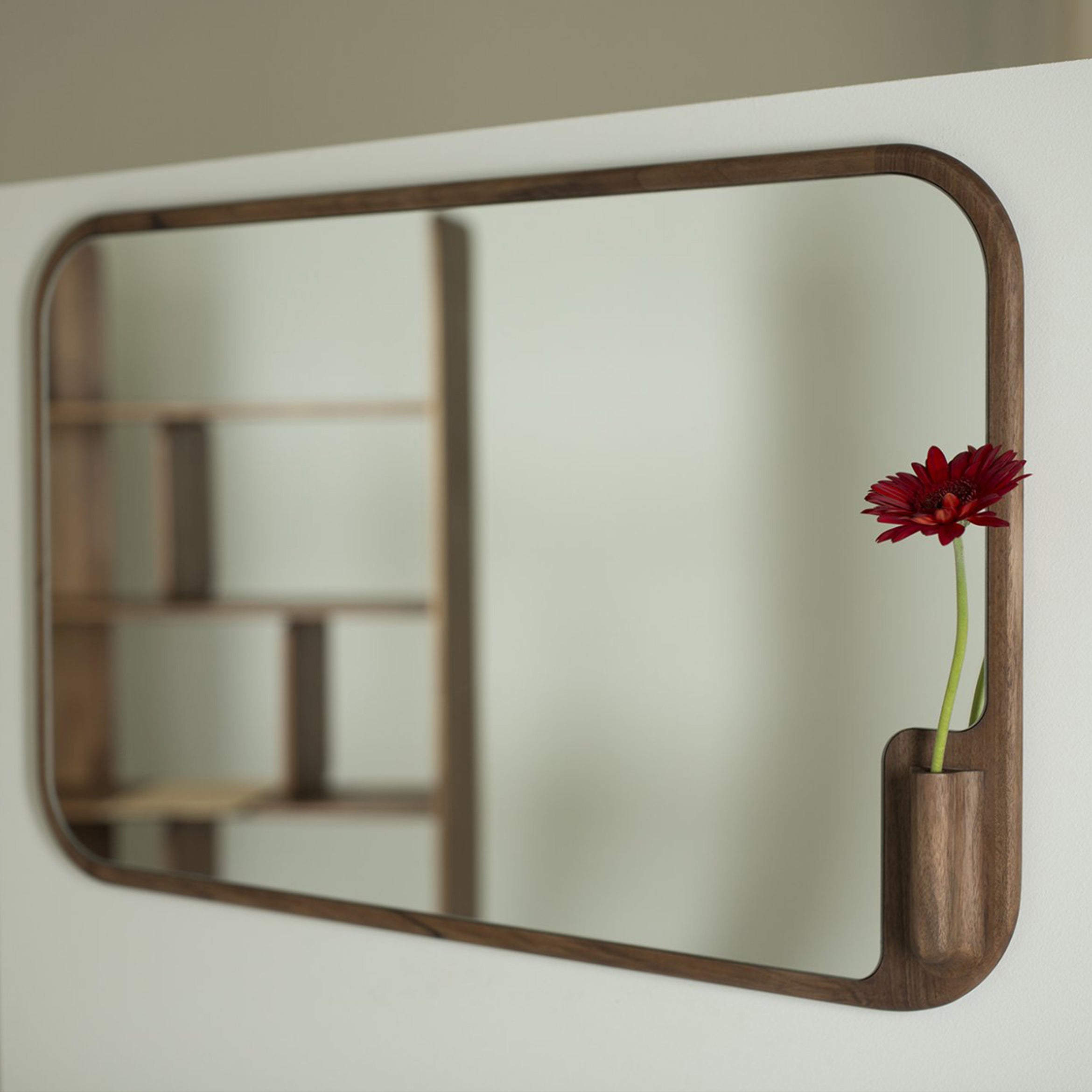 The MUSE Mirror by Artisan, a rectangular wood-framed piece with rounded corners, is mounted on a light wall. A narrow vase with a small red flower adorns its bottom right corner. Crafted from eco-friendly materials, it beautifully reflects shelving.