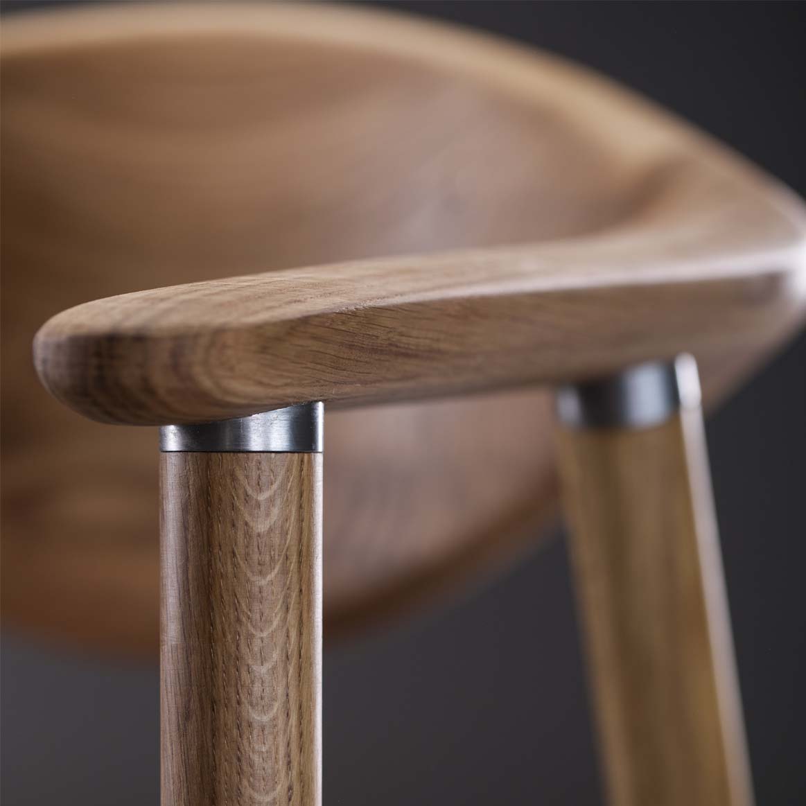 Close-up of the NARU Chair by Artisan, featuring an upholstered seat and backrest crafted from solid wood, with smooth curves and rounded armrests. Finished with natural oils and sleek metal accents at the joints. The softly out-of-focus dark background enhances its elegance.