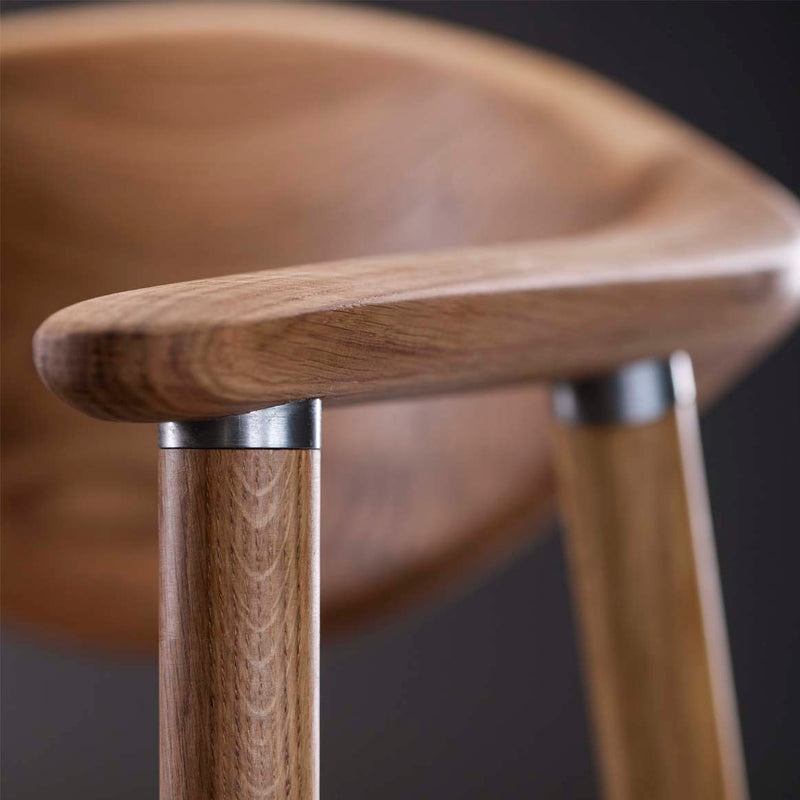 Close-up of the NARU Chair by Artisan, featuring an upholstered seat and backrest crafted from solid wood, with smooth curves and rounded armrests. Finished with natural oils and sleek metal accents at the joints. The softly out-of-focus dark background enhances its elegance.