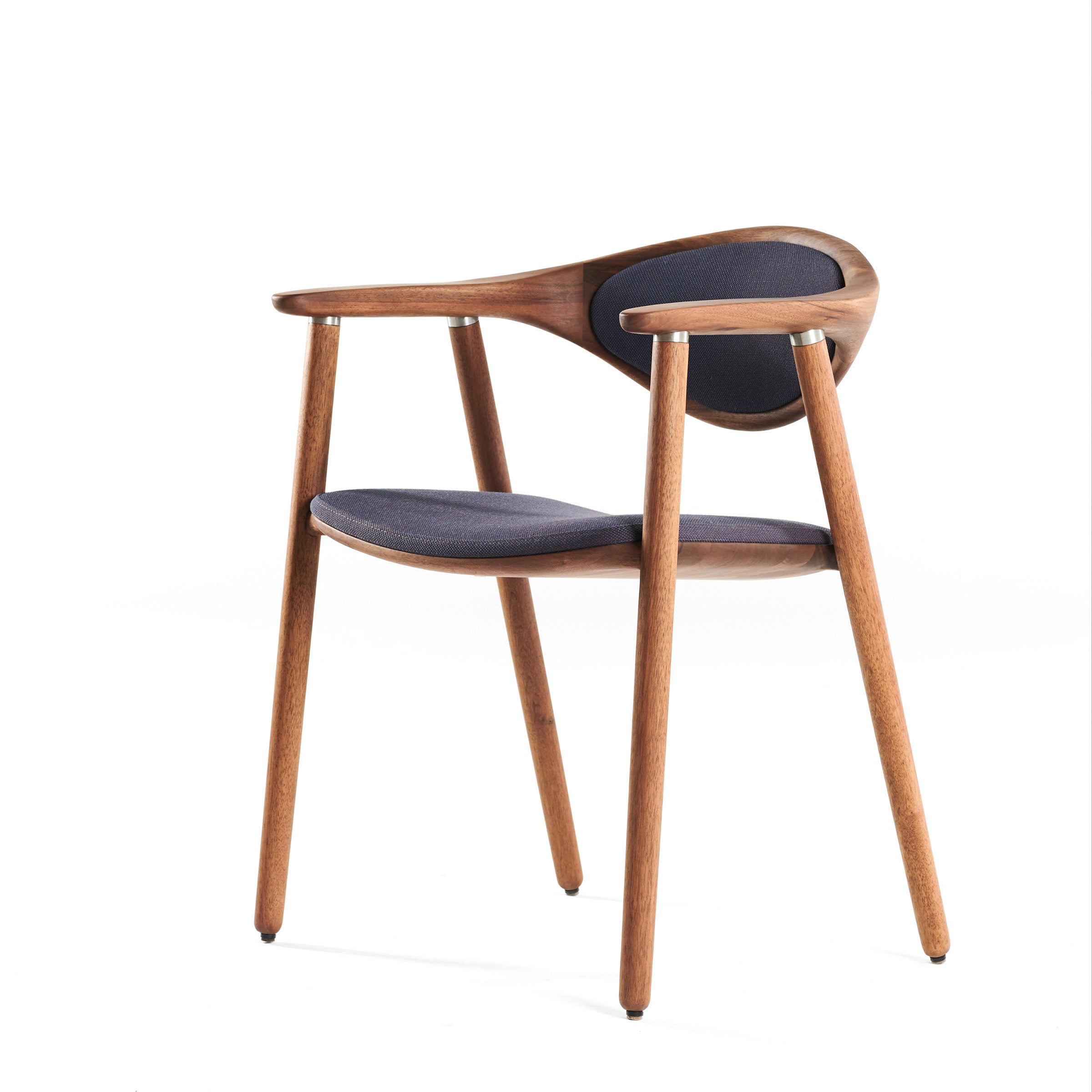 The Artisan NARU Chair is a modern solid wood chair with a sleek design. It features a curved backrest and armrests, upholstered in dark fabric. Finished with natural oils, it stands on four slender legs, showcasing a minimalist and contemporary style against a white background.
