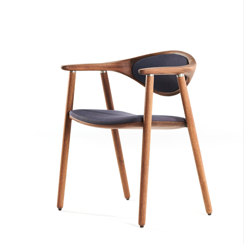 The Artisan NARU Chair is a modern solid wood chair with a sleek design. It features a curved backrest and armrests, upholstered in dark fabric. Finished with natural oils, it stands on four slender legs, showcasing a minimalist and contemporary style against a white background.