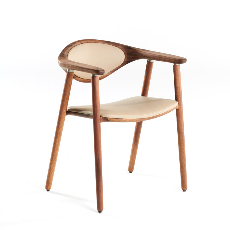 The Artisan NARU Chair is a modern design crafted from solid wood with a light beige upholstered seat and backrest. It features curved armrests, tapered legs, and is enhanced by natural oils, set against a plain white background.