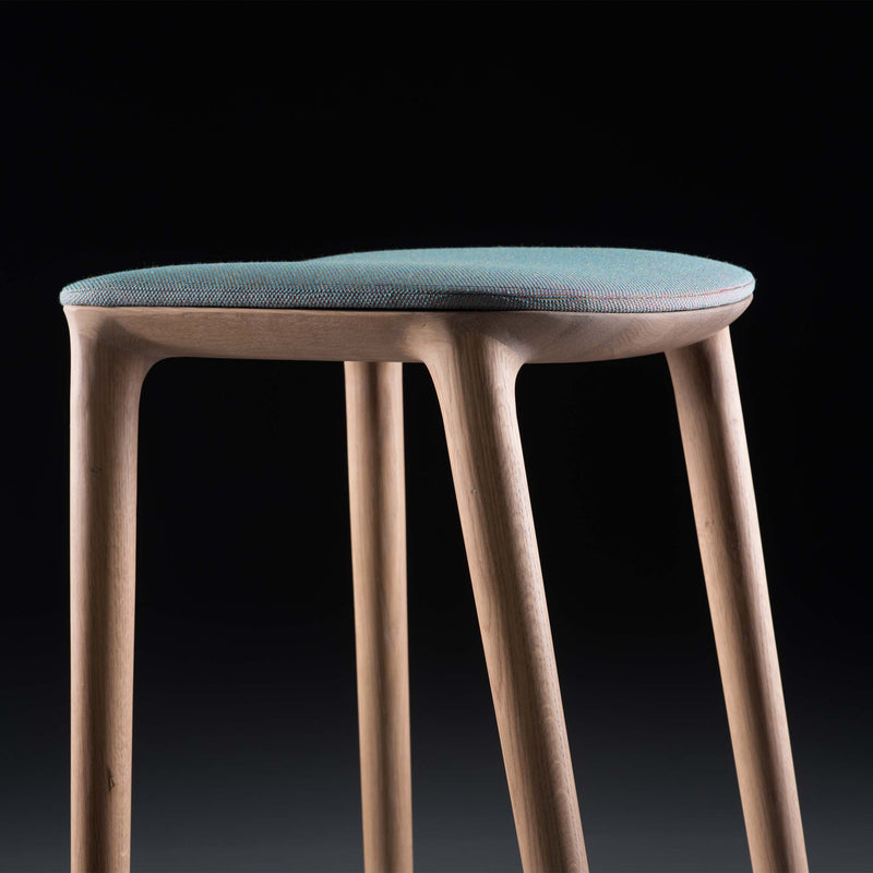 Close-up of the modern Artisan NEVA Stool upholstered, showcasing CNC-carved slender wooden legs and a blue cushioned seat, artfully set against a black background.