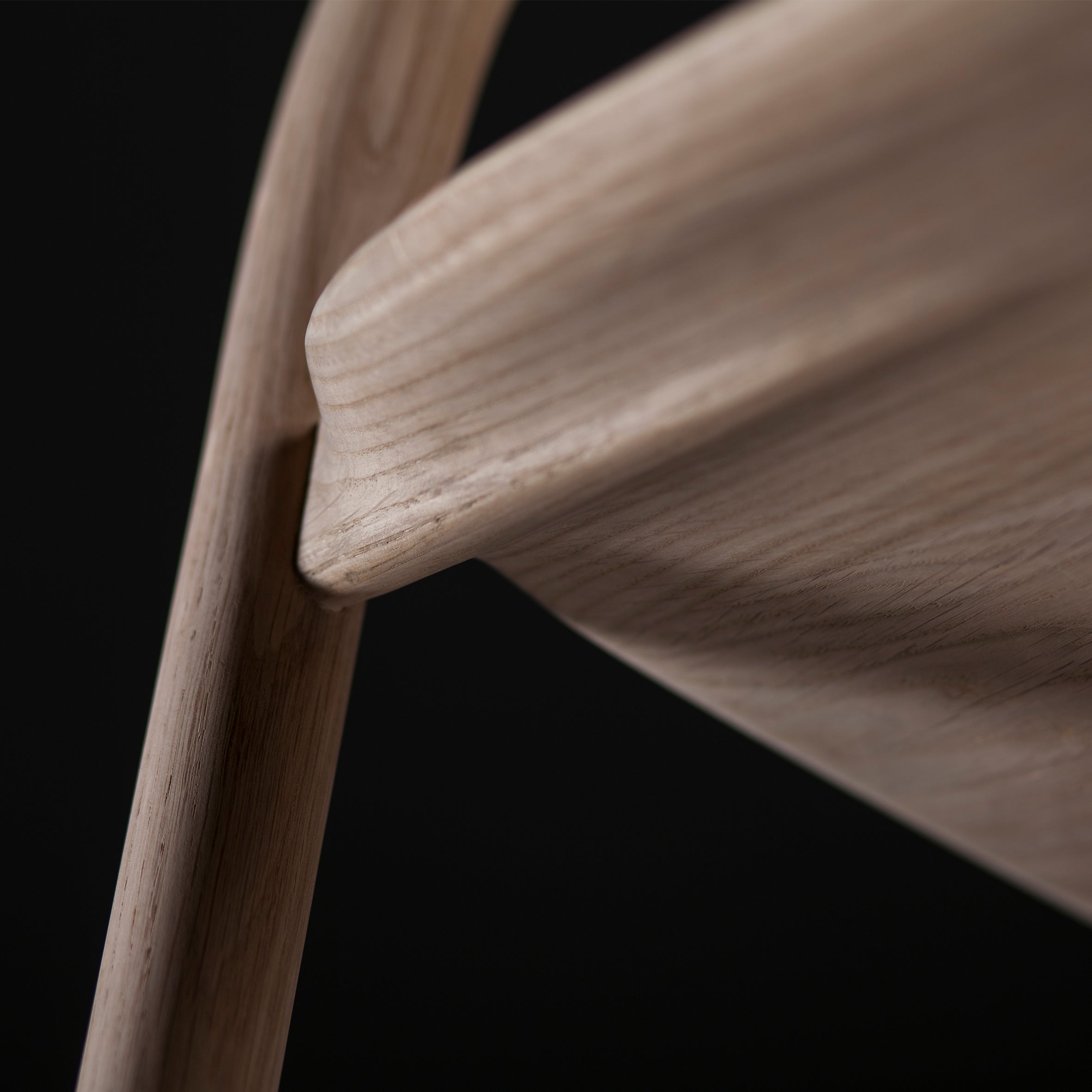 Close-up of an Artisan NEVA chair reveals the smooth, curved joint between the backrest and leg in American walnut. The light woods natural grain stands out against a dark background, enhanced by precise 5-axis CNC technology.