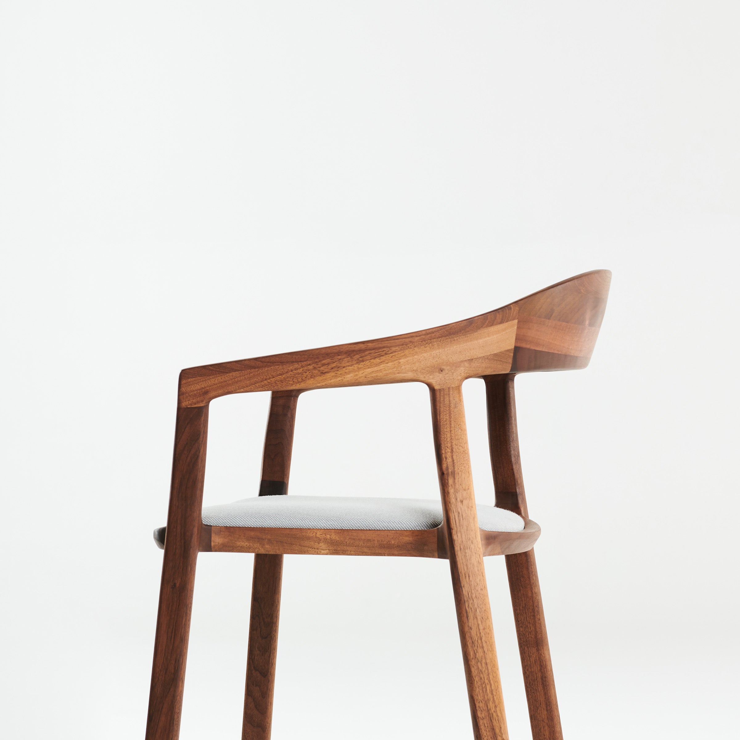 The TARA Armchair by Artisan is a modern wooden chair with a sleek design, featuring a curved backrest, smooth lines, and a light grey cushioned seat. Crafted from solid wood and finished with natural oils, its set against a plain white background.