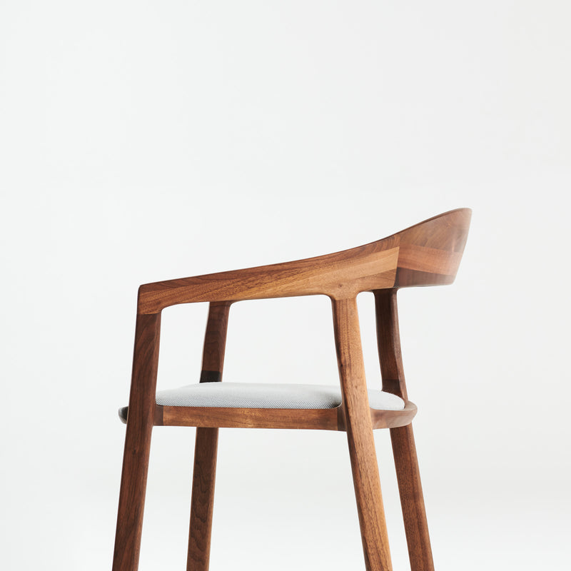 The TARA Armchair by Artisan is a modern wooden chair with a sleek design, featuring a curved backrest, smooth lines, and a light grey cushioned seat. Crafted from solid wood and finished with natural oils, its set against a plain white background.