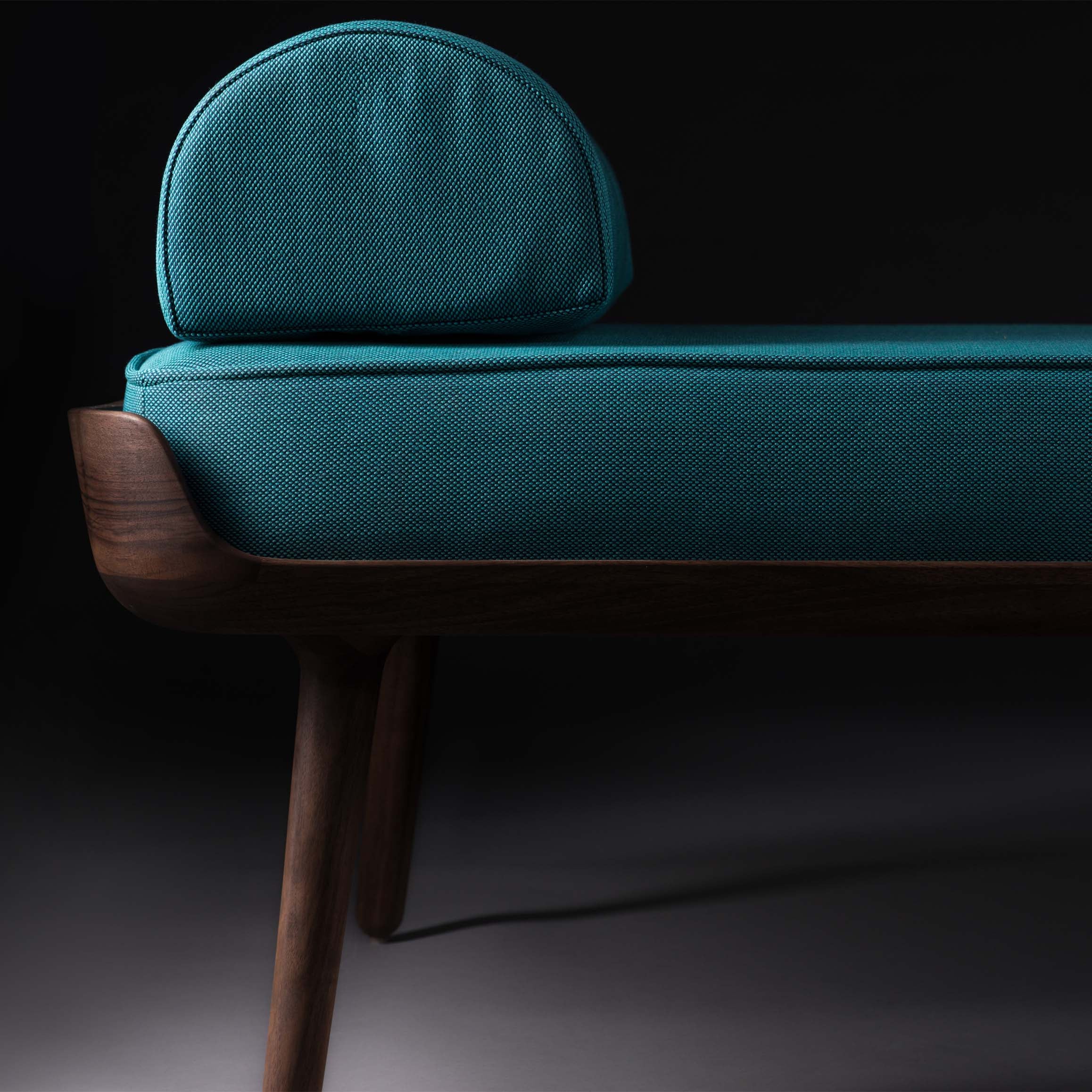 Close-up of the THOR Daybed by Artisan, featuring a sleek teal upholstered bench with natural wood-textured legs and a round bolster pillow. This modern design is set against a dark backdrop and crafted from environmentally-friendly materials.