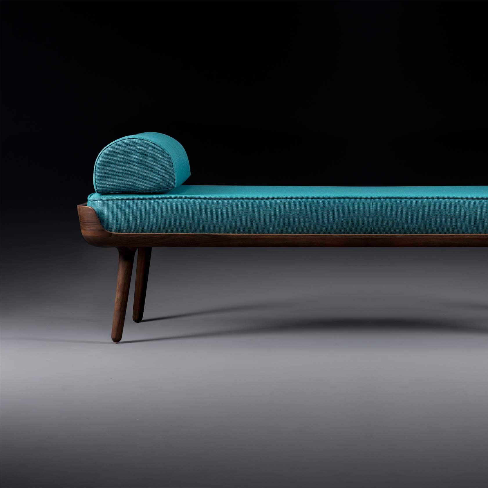 The Artisan THOR Daybed features teal upholstery, natural wood-textured wooden legs, and a cylindrical cushion at one end, set against a dark background.