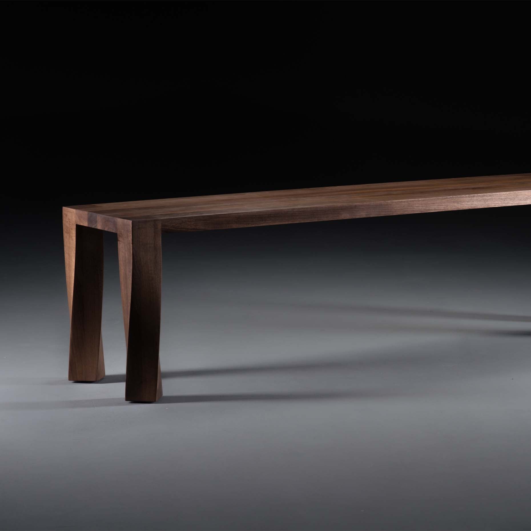 The TOR Bench by Artisan is a sleek, modern piece crafted from solid wood with a dark finish. It features clean lines and uniquely angled legs, set against a dark gray background, emphasizing its eco-friendly design enhanced by natural oils.