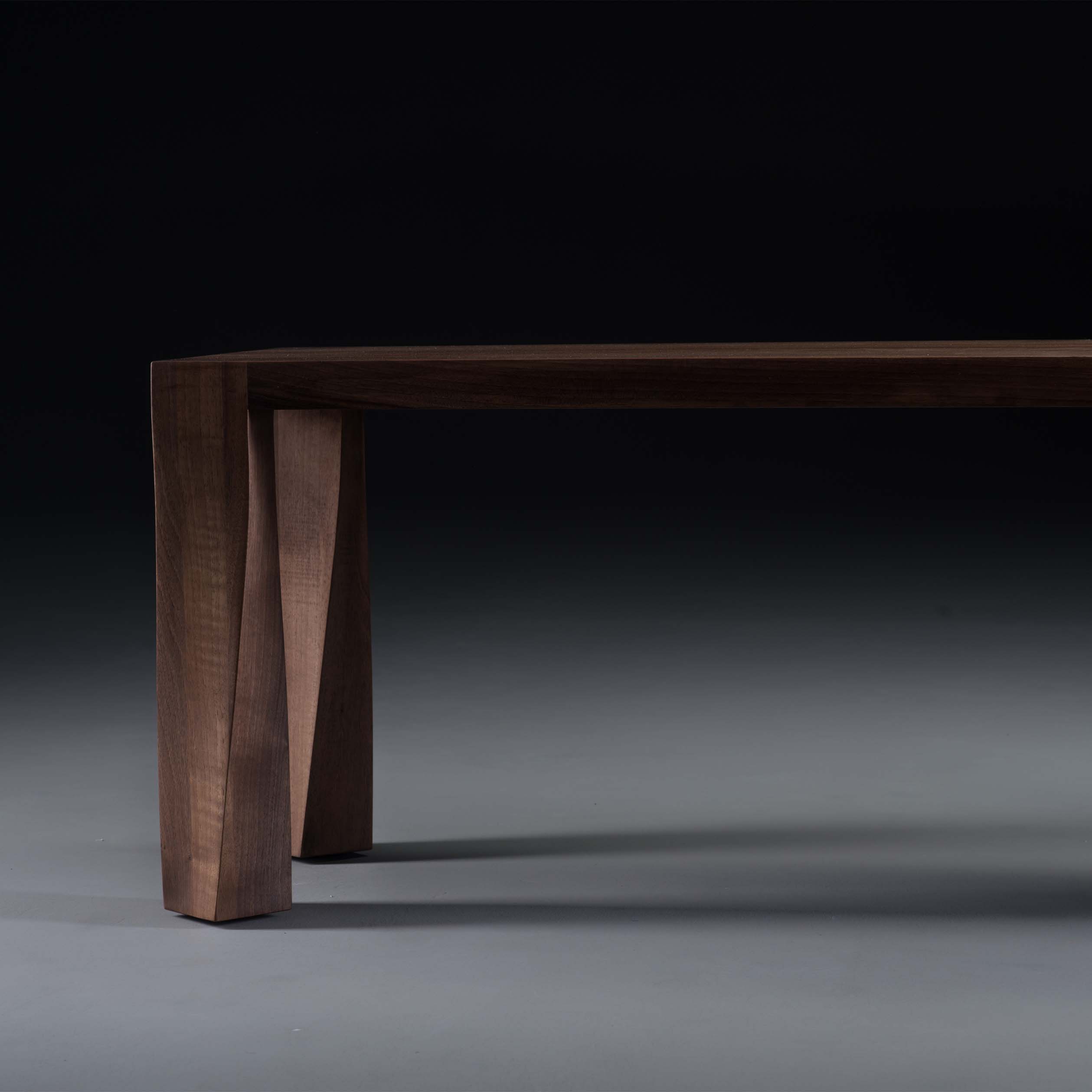 Close-up of the TOR Bench by Artisan, featuring a dark wooden tabletop and distinctive angled legs against a dark background. Crafted from solid wood and treated with natural oils, it highlights the wood grain for an eco-friendly, minimalist, modern look.