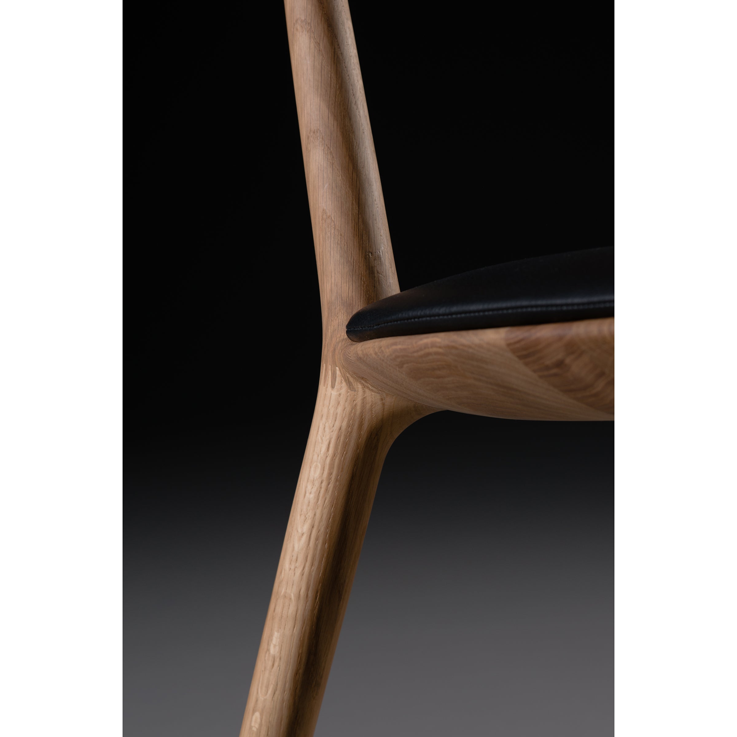 Close-up of the Artisan WU Chair Upholstered shows a solid wood frame with a natural finish, enhanced by oils. Its black seat contrasts with the light wood, highlighting joinery connecting the leg to the seat against a dark background.