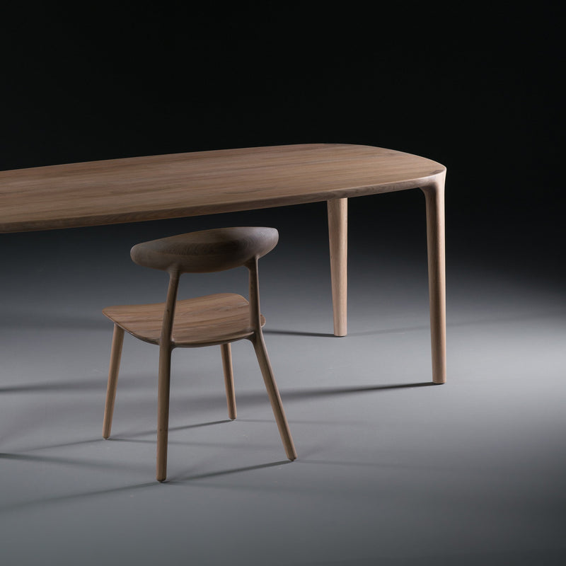 Set against a dark background, the Artisan WU Table pairs with a minimalist wooden chair. Crafted from solid wood, the chair features a curved backrest and slender legs, highlighting the luxurious texture of the tables smooth, rounded edges.