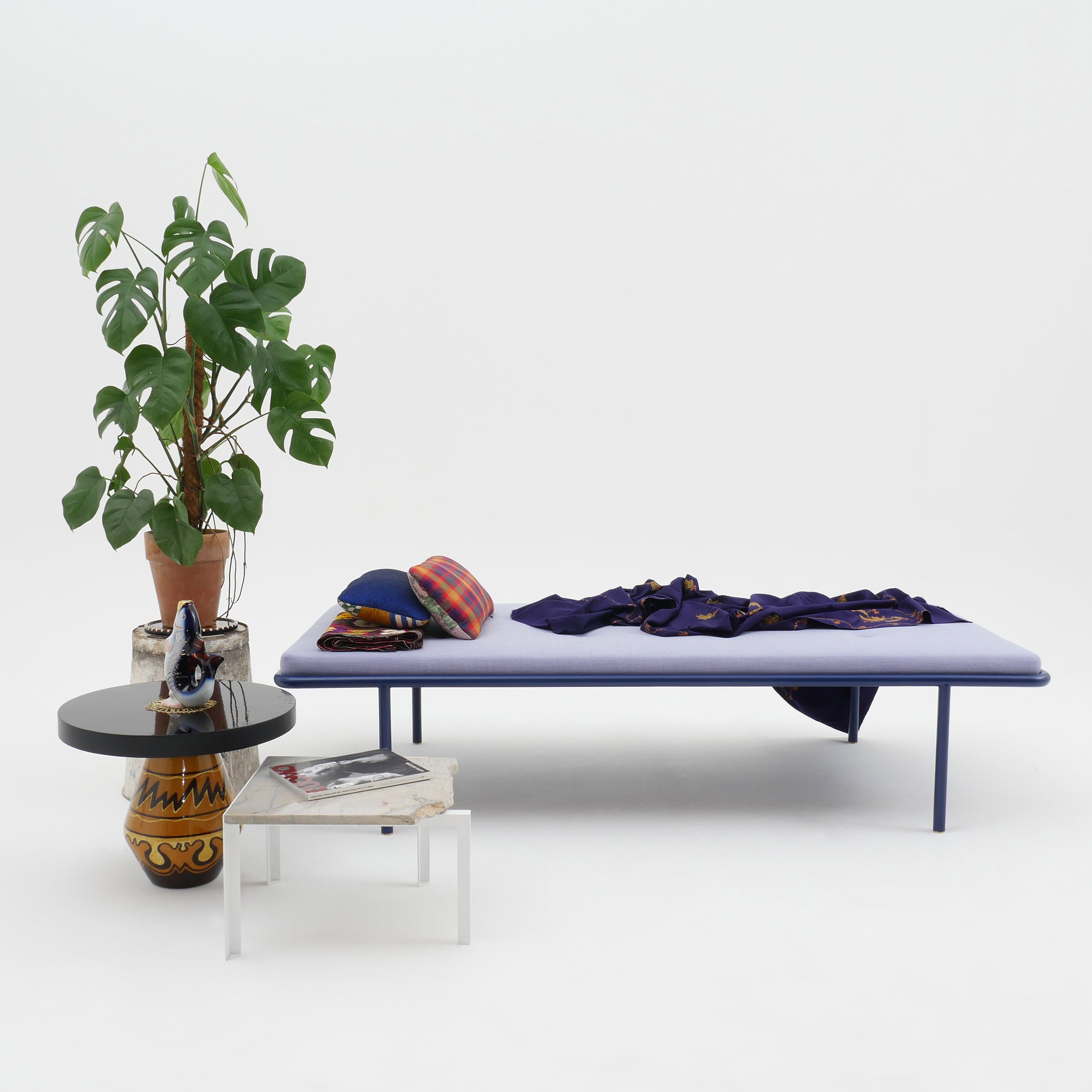 2PM Day & Night Daybed with blue frame and cushion, shown with side table and decorative monstera plant
