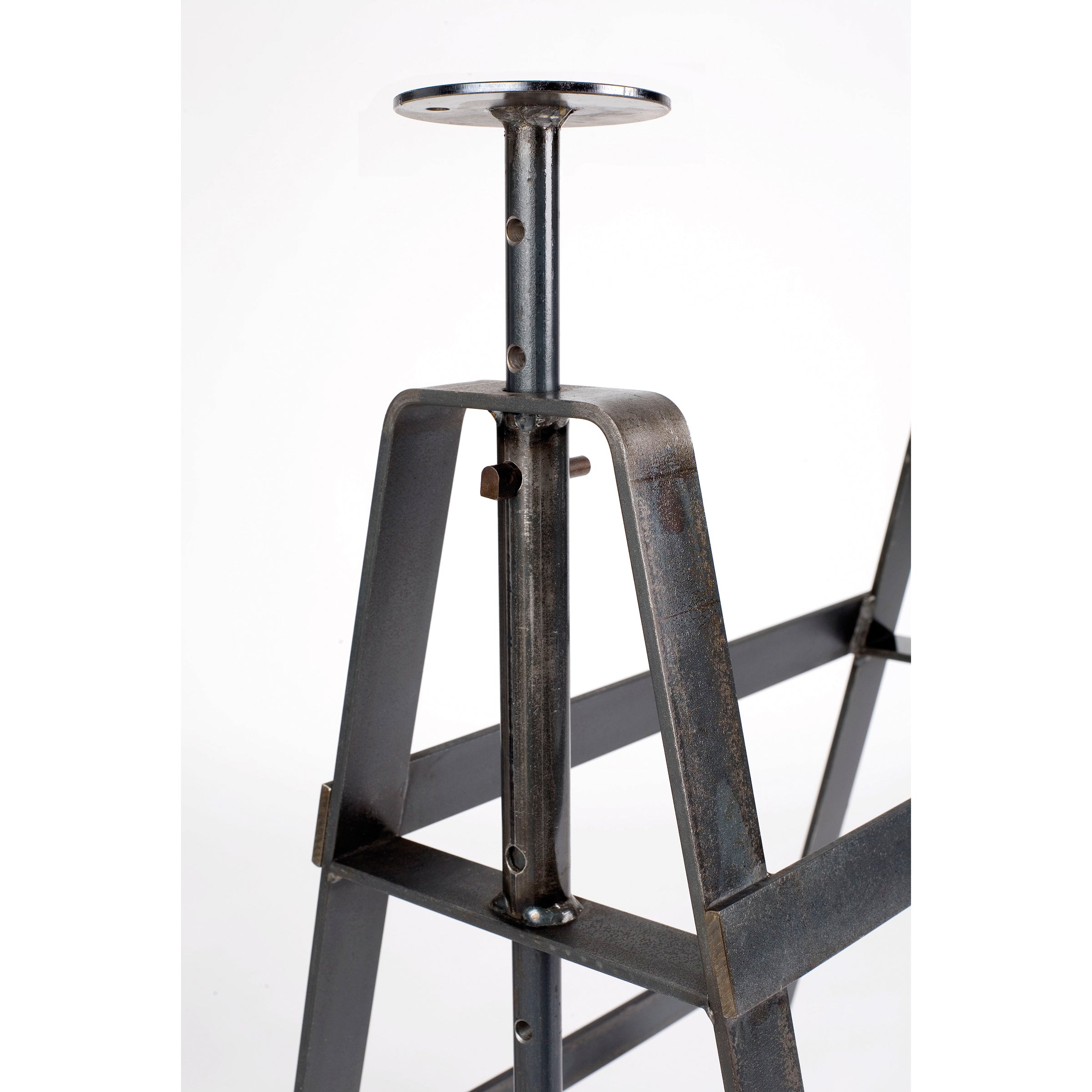 Close-up of the Affe Trestle by Atelier Haussmann, an industrial-style, height-adjustable table base in thick, dark oiled raw steel with visible screws and a vertical adjustment mechanism, topped with a round plate. The plain white background highlights its rugged elegance.