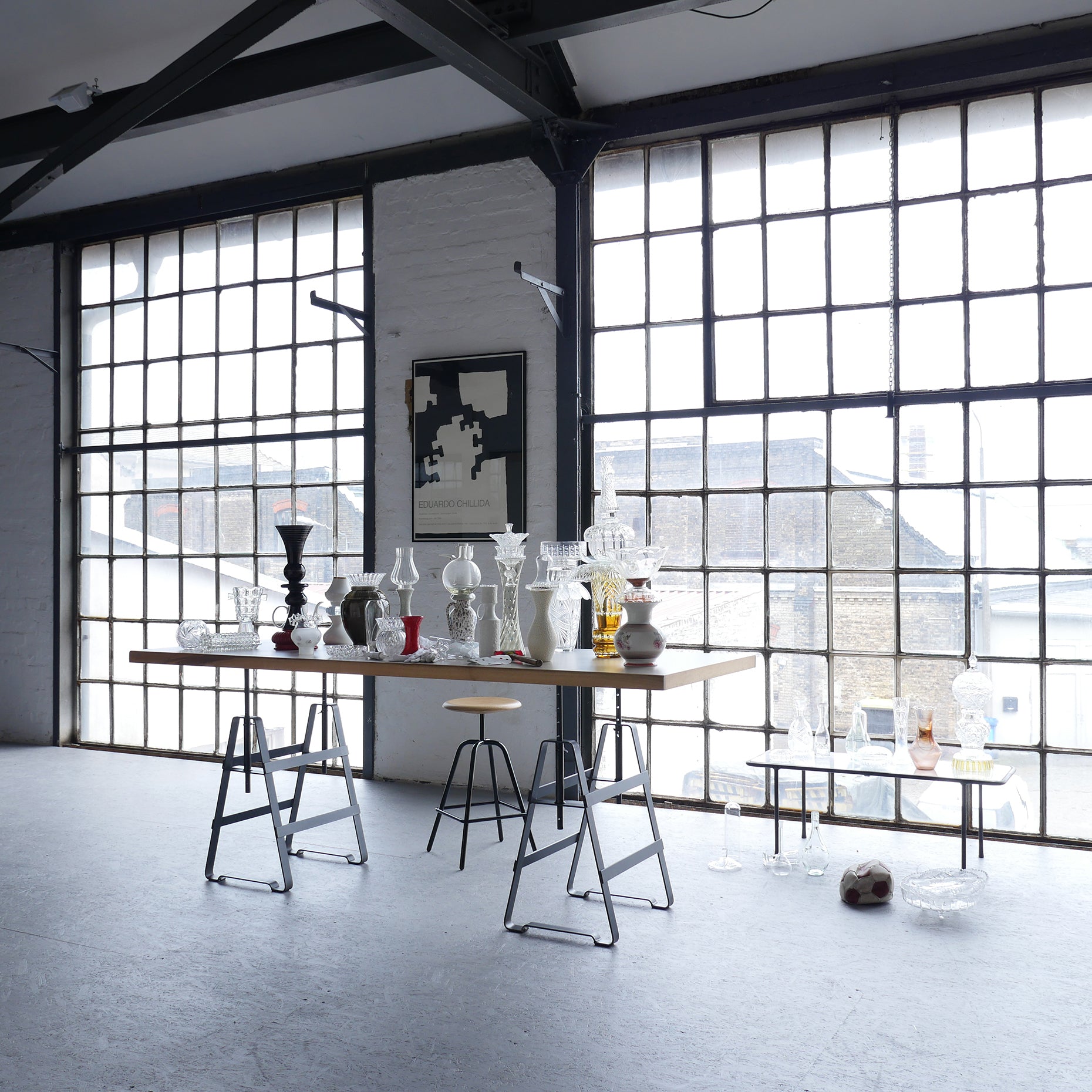 A spacious studio with large industrial windows features an Affe Trestle table by Atelier Haussmann displaying glass vases and sculptures. An abstract wall poster and a Berlin-crafted stool highlight the minimalist space, bathed in natural light.