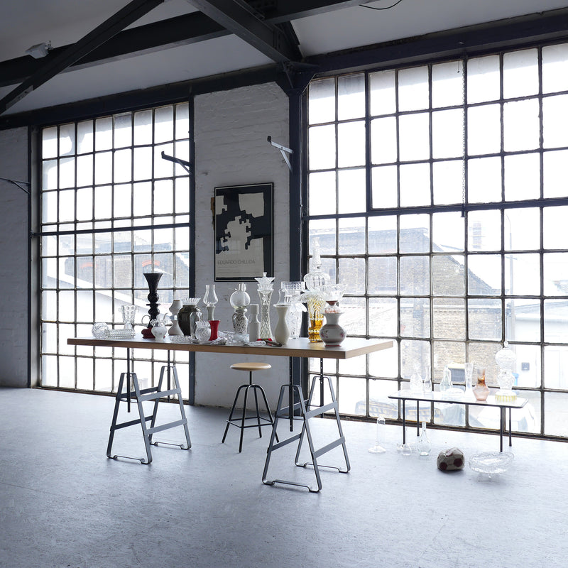 A spacious studio with large industrial windows features an Affe Trestle table by Atelier Haussmann displaying glass vases and sculptures. An abstract wall poster and a Berlin-crafted stool highlight the minimalist space, bathed in natural light.