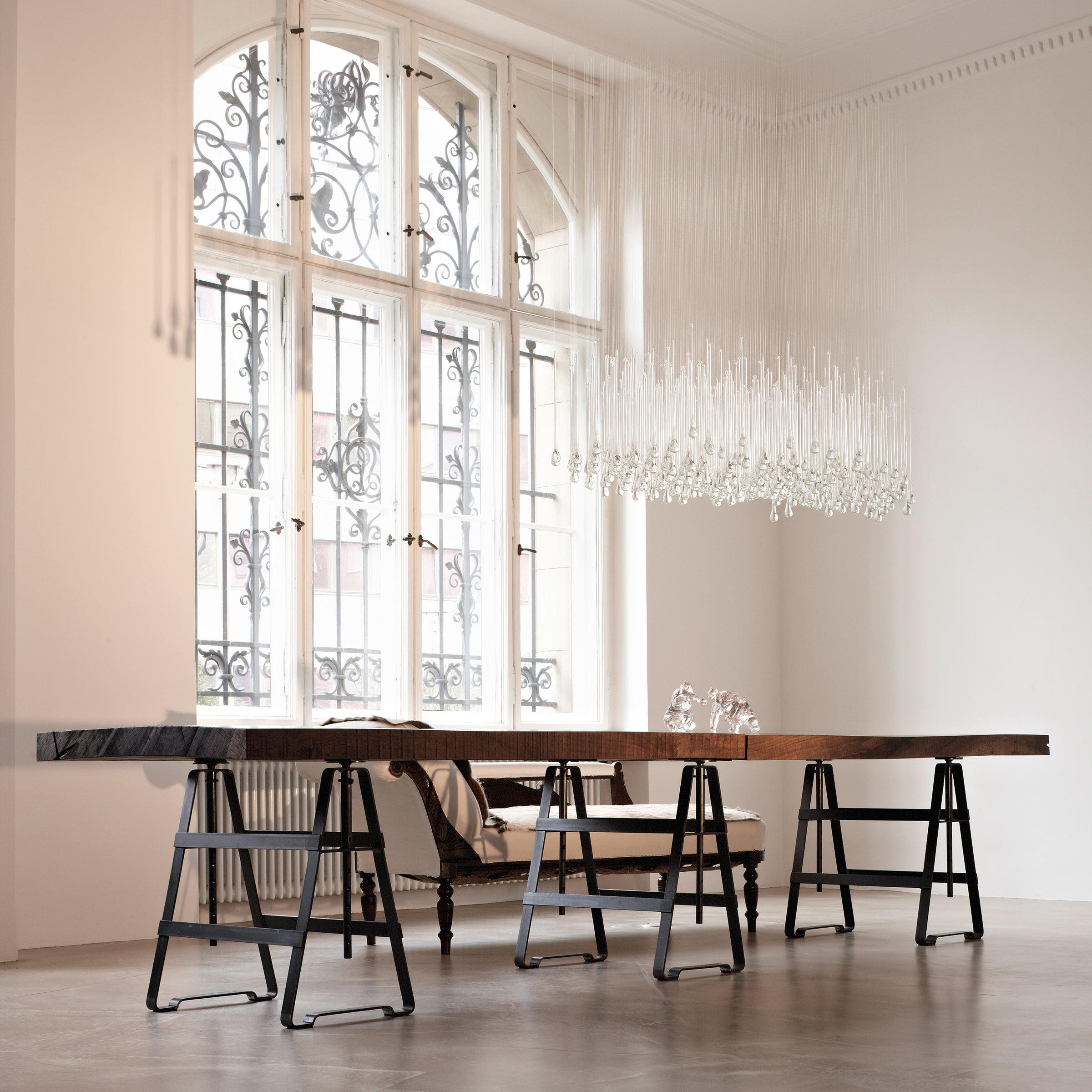 In a minimalist dining room, an Affe Trestle table by Atelier Haussmann showcases height-adjustable oiled raw steel legs under a modern chandelier of hanging glass rods. Large windows with ornate ironwork fill the space with light, emphasizing a Berlin craft producers minimalist aesthetic.