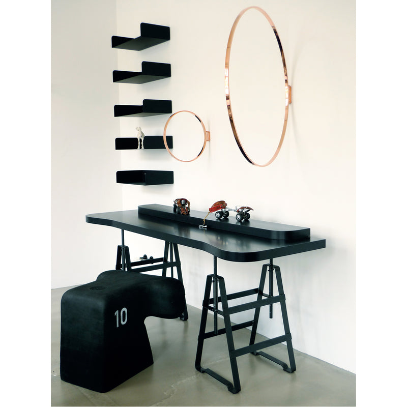 A modern workspace showcases the sleek black Affe Trestle desk by Atelier Haussmann, with industrial-style oiled raw steel legs. Decorative items, circular mirrors, a black wall shelf, and a sculpted 10 stool complete the contemporary look.