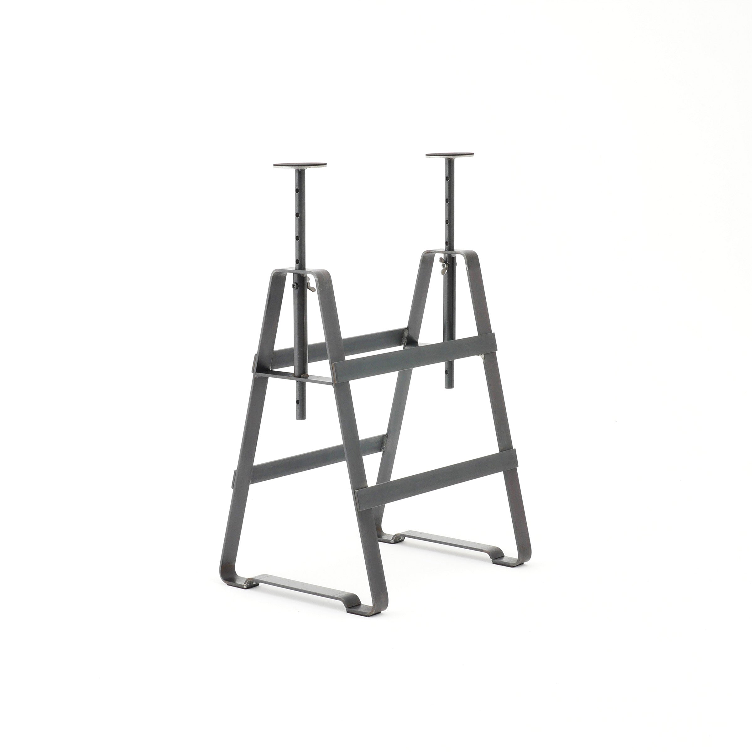 The Affe Trestle by Atelier Haussmann is a gray metal saw horse with adjustable height trestle legs, two A-frame ends, and a horizontal support beam. Its simple yet functional design in oiled raw steel is crafted by Berlin artisans against a bright background.
