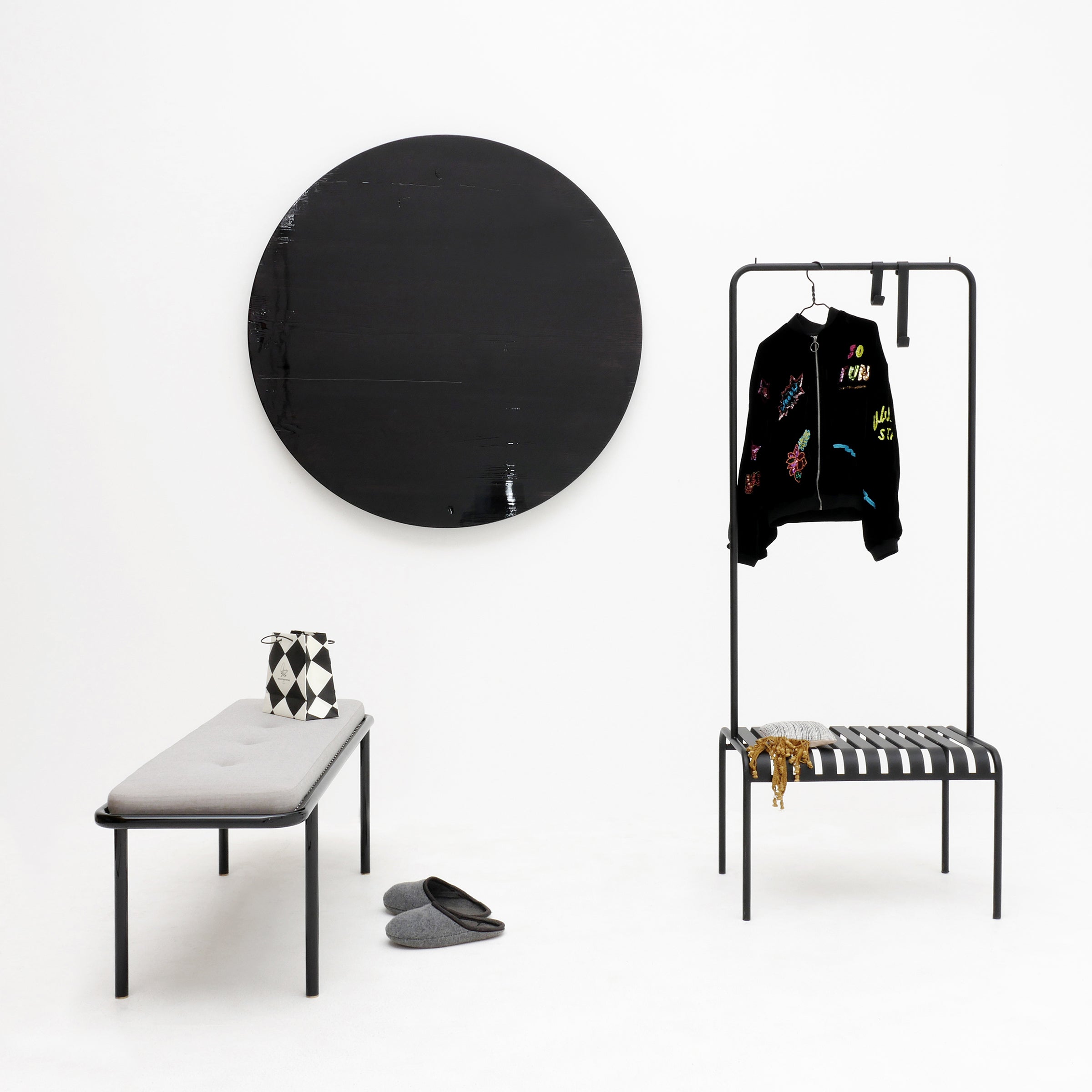 A minimalist room showcases a black circular wall art and a gray bench with a patterned pillow. An Atelier Haussmann YOUTOO clothing rack in powder-coated steel holds a black jacket with colorful designs, while gray slippers rest on the floor, enhancing the modern monochrome aesthetic.