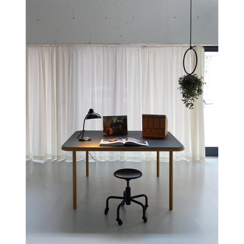 A minimalist workspace has a table and black stool. The table holds a lamp, book, magazine, and stylish file holders. Sheer curtains hang behind with an Atelier Haussmann Blumenkugel hanging to the side as natural light filters in.