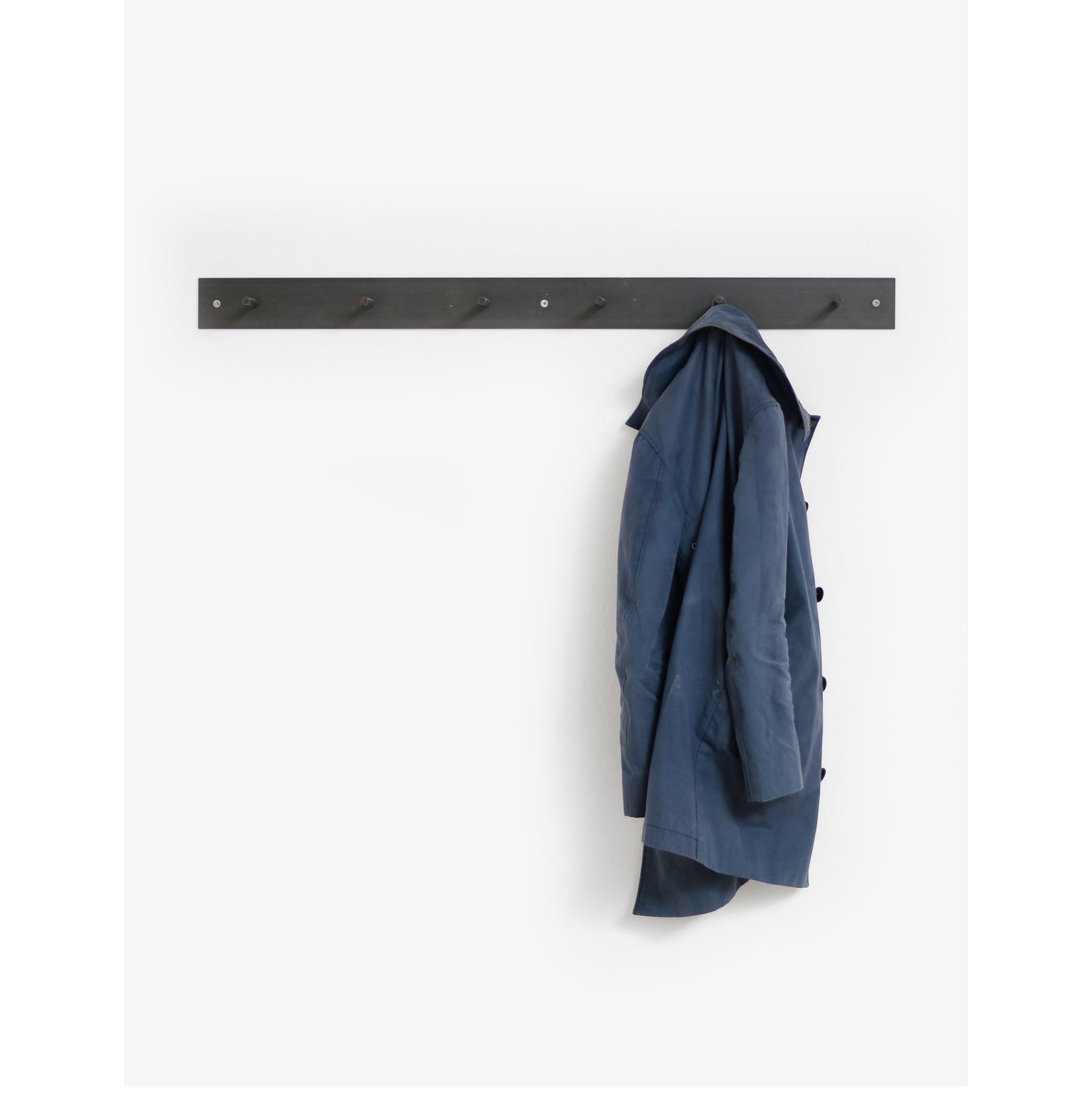 A long, dark wooden Atelier Haussmann HELLOGOODBYE coatrack with multiple pegs is mounted on a white wall. A blue trench coat hangs from one peg, while the rest are empty.