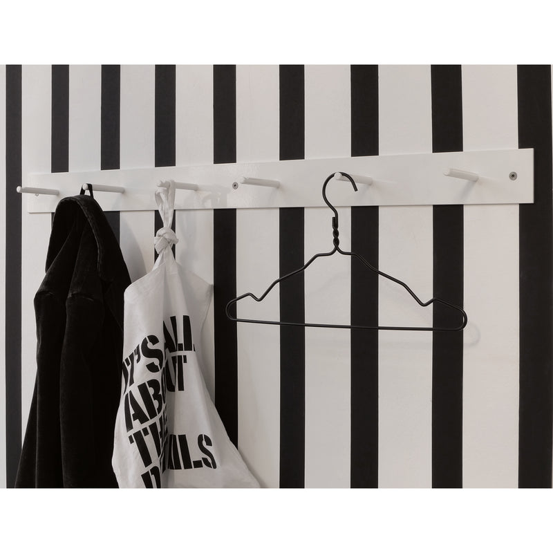 A black and white striped wall showcases an Atelier Haussmann HELLOGOODBYE coatrack made of white powder-coated steel, elegantly holding a black coat, a text-printed white cloth bag, and an empty wire hanger.