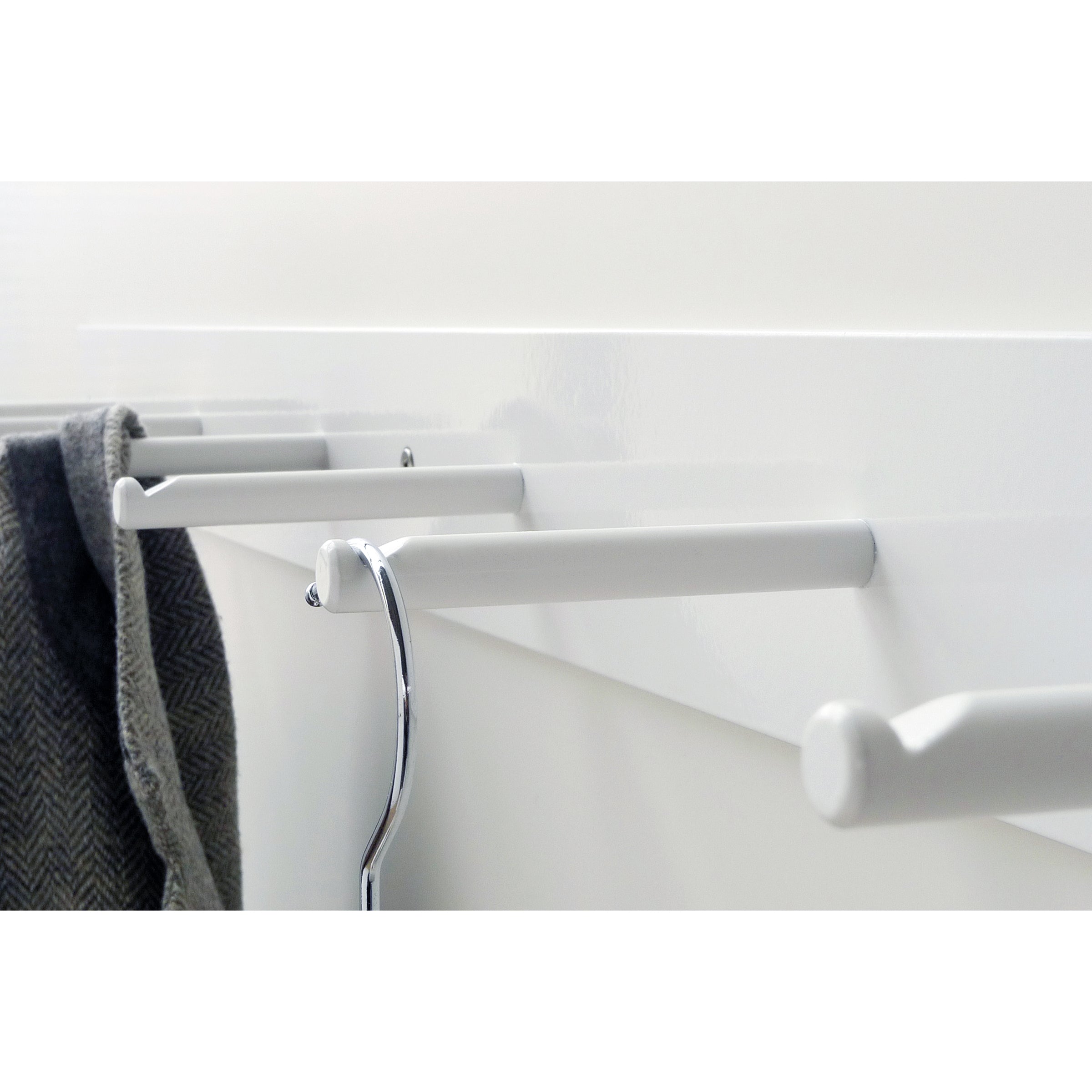 A close-up of Atelier Haussmann’s HELLOGOODBYE wall-mounted coatrack, made from white powder-coated steel, shows one metal hanger on a peg and a dark, textured fabric draped over another, capturing the linear arrangement of pegs beautifully.