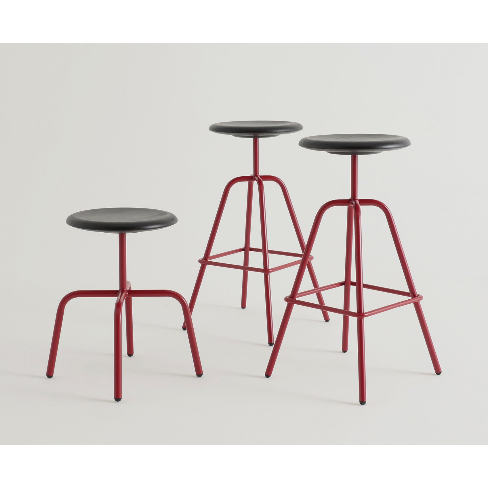 Three Herrenberger stools by Atelier Haussmann, featuring black round seats and red metal legs, stand against a plain white background. Each stool varies slightly in height, displaying a simple, minimalist design with an industrial appeal.