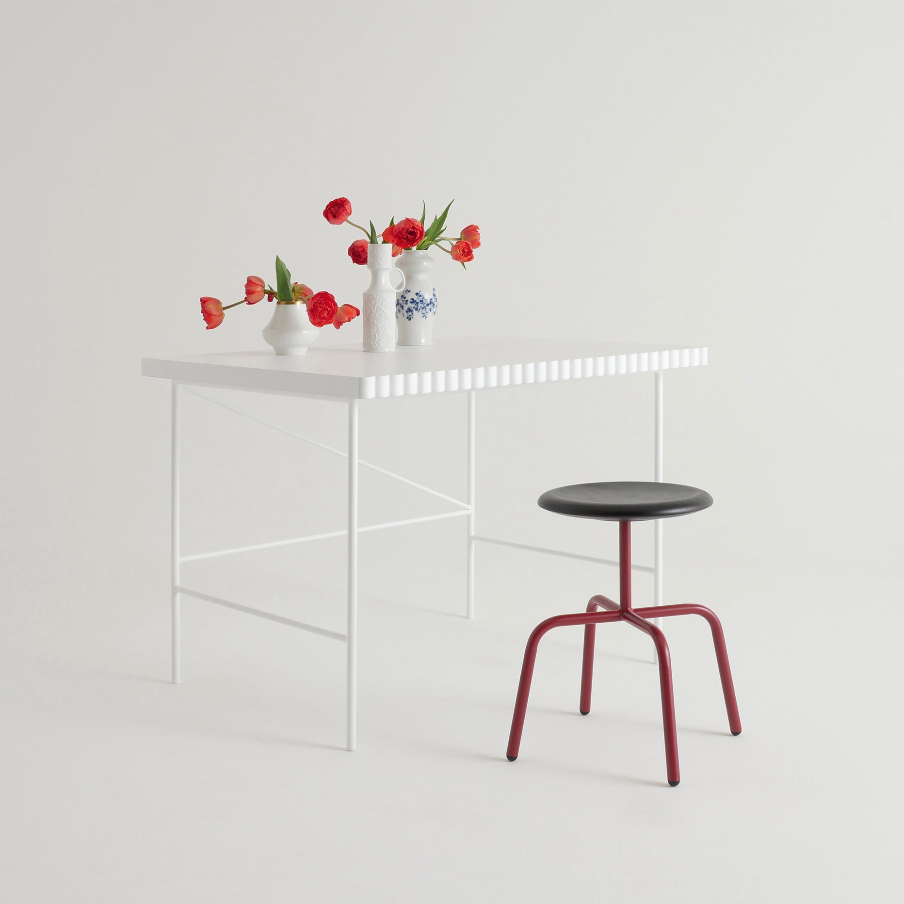 A minimalist scene features a white table with a striped-edge design and three vases of red flowers. Beside it sits an Atelier Haussmann Herrenberger stool, showcasing red legs, a black seat, and a tubular steel frame for an industrial touch against the plain white backdrop.