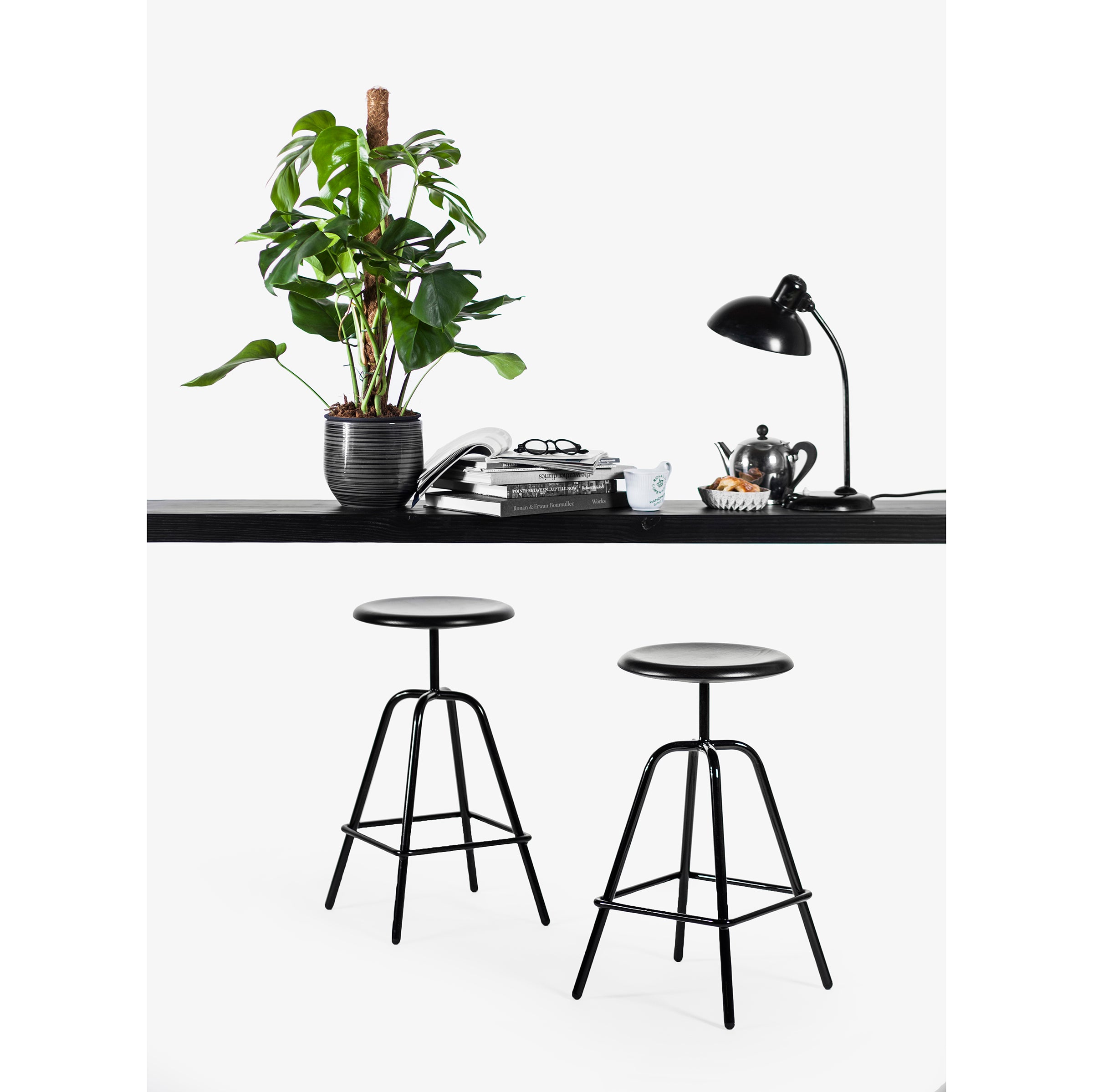 A modern workspace features a black desk with a potted plant, books, and a sleek lamp. Beneath, two Herrenberger Barstools by Atelier Haussmann, featuring circular seats and tubular steel frames, stand against a white backdrop epitomizing industrial design.