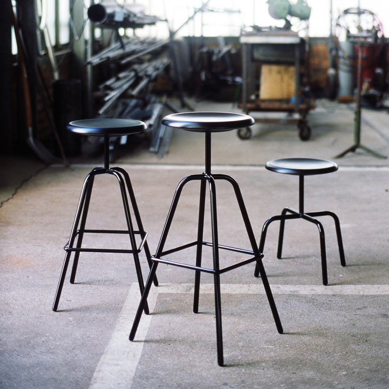 Three Herrenberger Barstools by Atelier Haussmann, with varying heights and sleek tubular steel frames, are set on a concrete workshop floor. Blurred equipment in the background enhances the industrial vibe, while bright lighting accentuates their modern elegance.