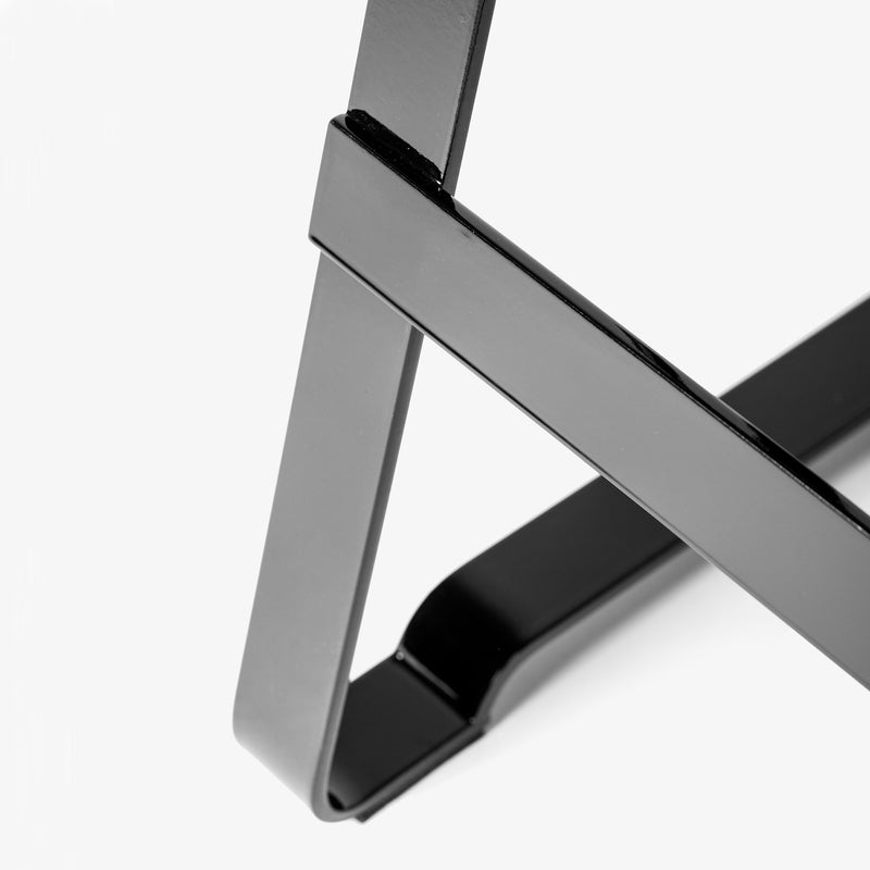 Close-up of Atelier Haussmanns Lackaffe Trestle, a sleek black metal stand made by Berlins artisans. It features intersecting geometric shapes on a white background, showcasing smooth, shiny powder-coated steel surfaces and sharp angles in a minimalist design.