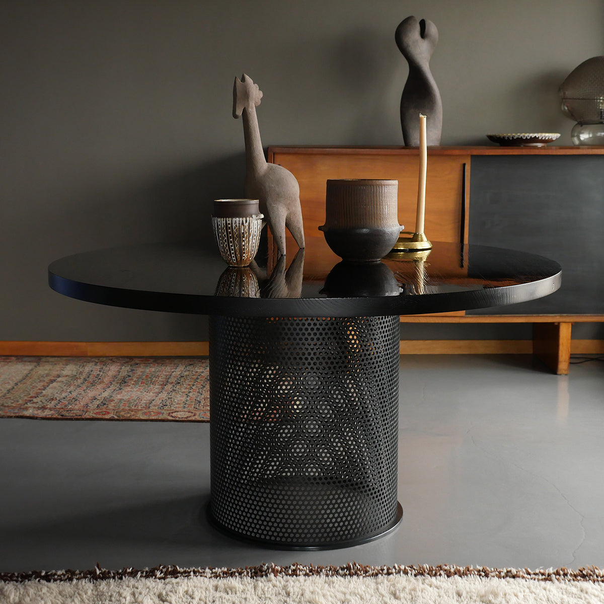 A modern living room showcases the Leipzig Table by Atelier Haussmann, featuring an ash wood tabletop and perforated base. It holds decorative items like a giraffe figurine, candle holder, and small vase. Completing the minimalist look are a wooden sideboard and a textured rug.