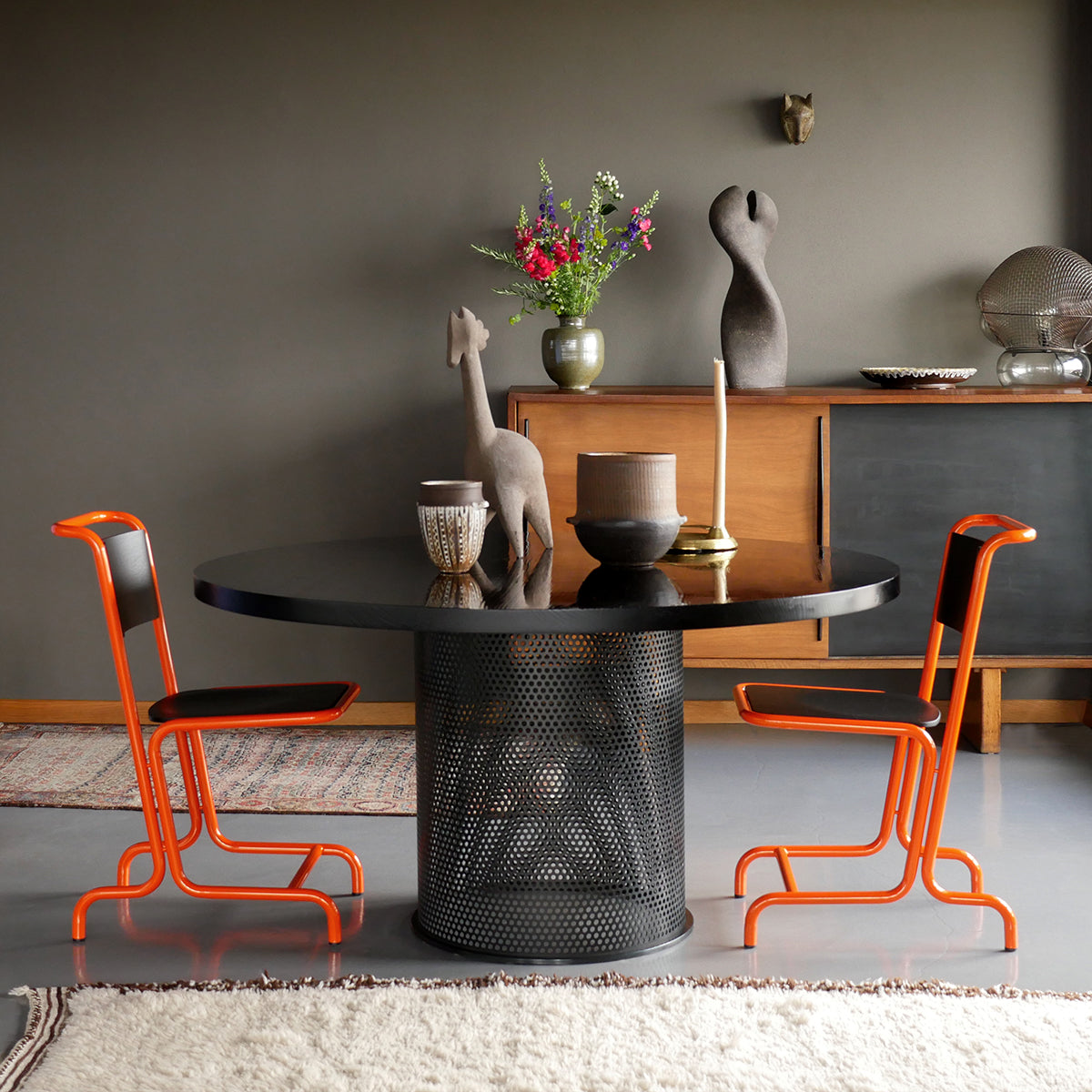 A modern dining room showcases an Atelier Haussmann Leipzig Table with an ash wood tabletop, paired with two vibrant orange chairs. A giraffe sculpture and vase enhance the decor, a wooden sideboard rests against gray walls, and a light rug unifies the space.