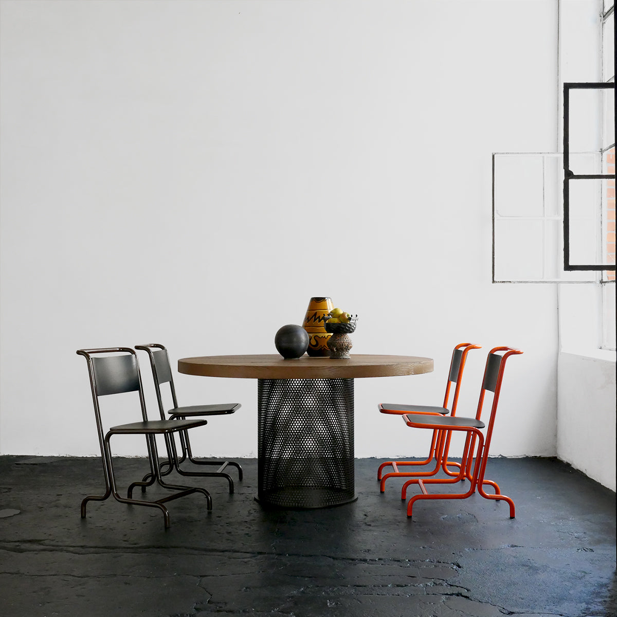 A modern dining area features Atelier Haussmanns Leipzig Table with an ash wood top and a metal base. It is surrounded by two black and two orange metal chairs. Decorative vases and a plant enhance the space, while large windows allow in ample natural light.