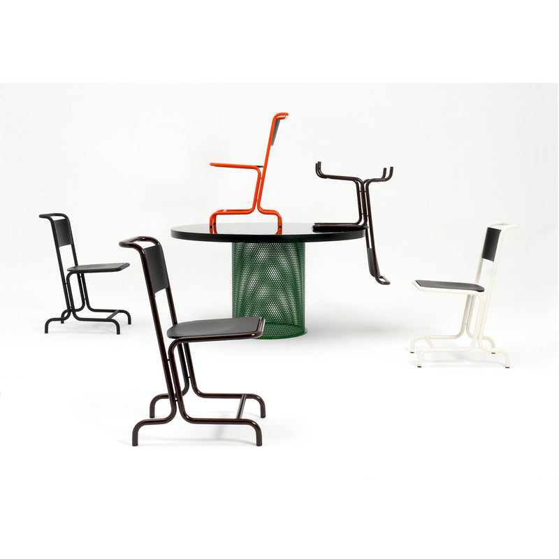 Four modern minimalist chairs in black, white, and red encircle and rest on a black circular Leipzig Table by Atelier Haussmann, featuring a green perforated steel sheet base. The arrangement stands against a plain white backdrop, highlighting the designs creativity with curved legs.