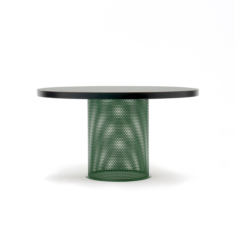 The Atelier Haussmann Leipzig Table features a sleek black or ash wood top, a unique green perforated steel cylindrical base with a geometric pattern, and is set against a plain white background.