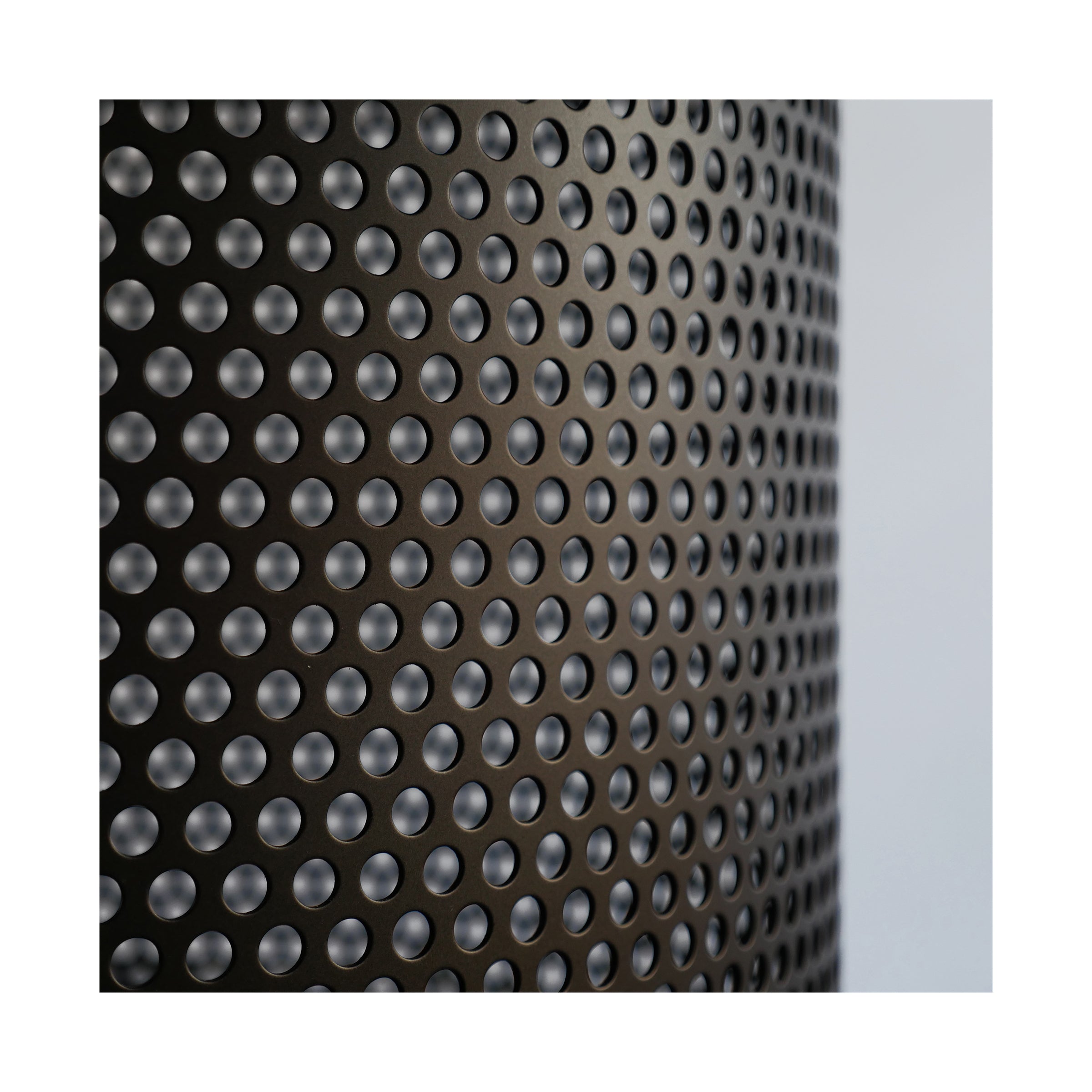 Close-up of the Leipzig Table by Atelier Haussmann, showcasing a sleek cylindrical shape with a perforated steel sheet surface and evenly spaced circular holes. The lighting enhances its metallic texture for a modern, industrial aesthetic against a softly blurred background.