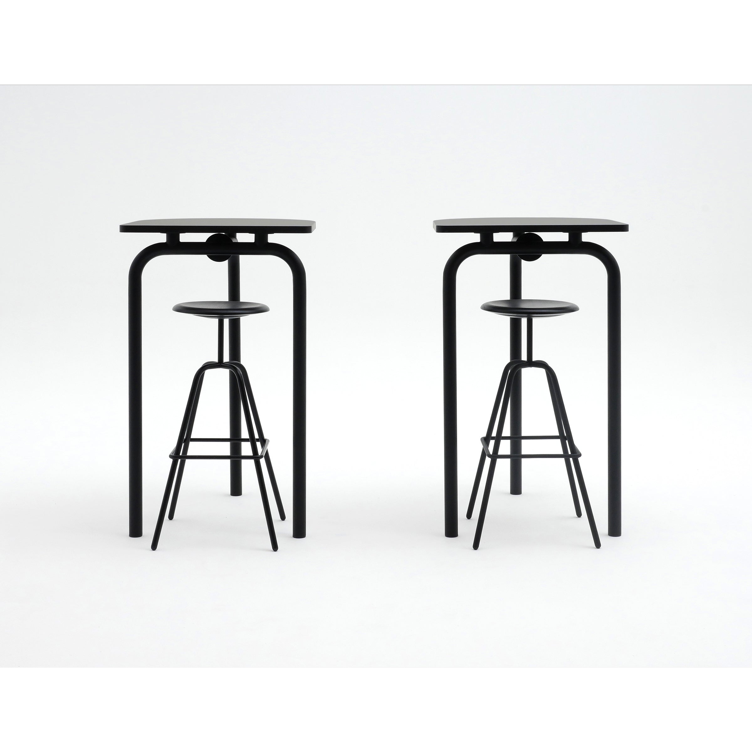 Two sleek black bar stools are set in front of matching Piombino Alto 700 tables by Atelier Haussmann, highlighting minimalist and industrial design. Each stool has a circular seat and narrow legs, while the tables feature rectangular surfaces with four slender legs.