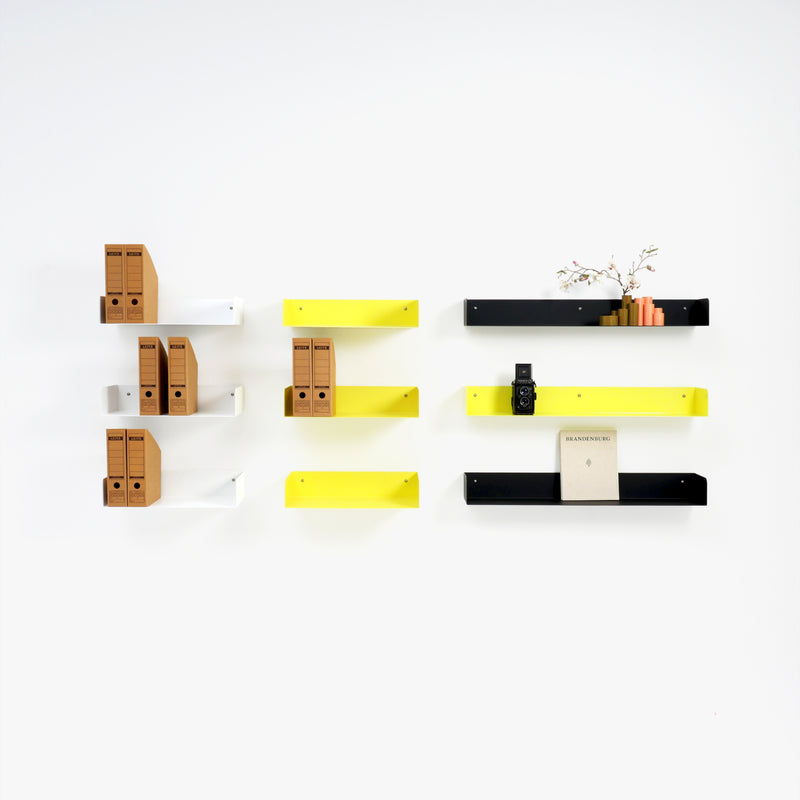 A minimalist wall showcases Atelier Haussmanns Poggibonsi steel shelves in white, yellow, and black. The powder-coated shelves neatly display brown file boxes, a digital clock, a book, and a small plant against the pristine white backdrop.