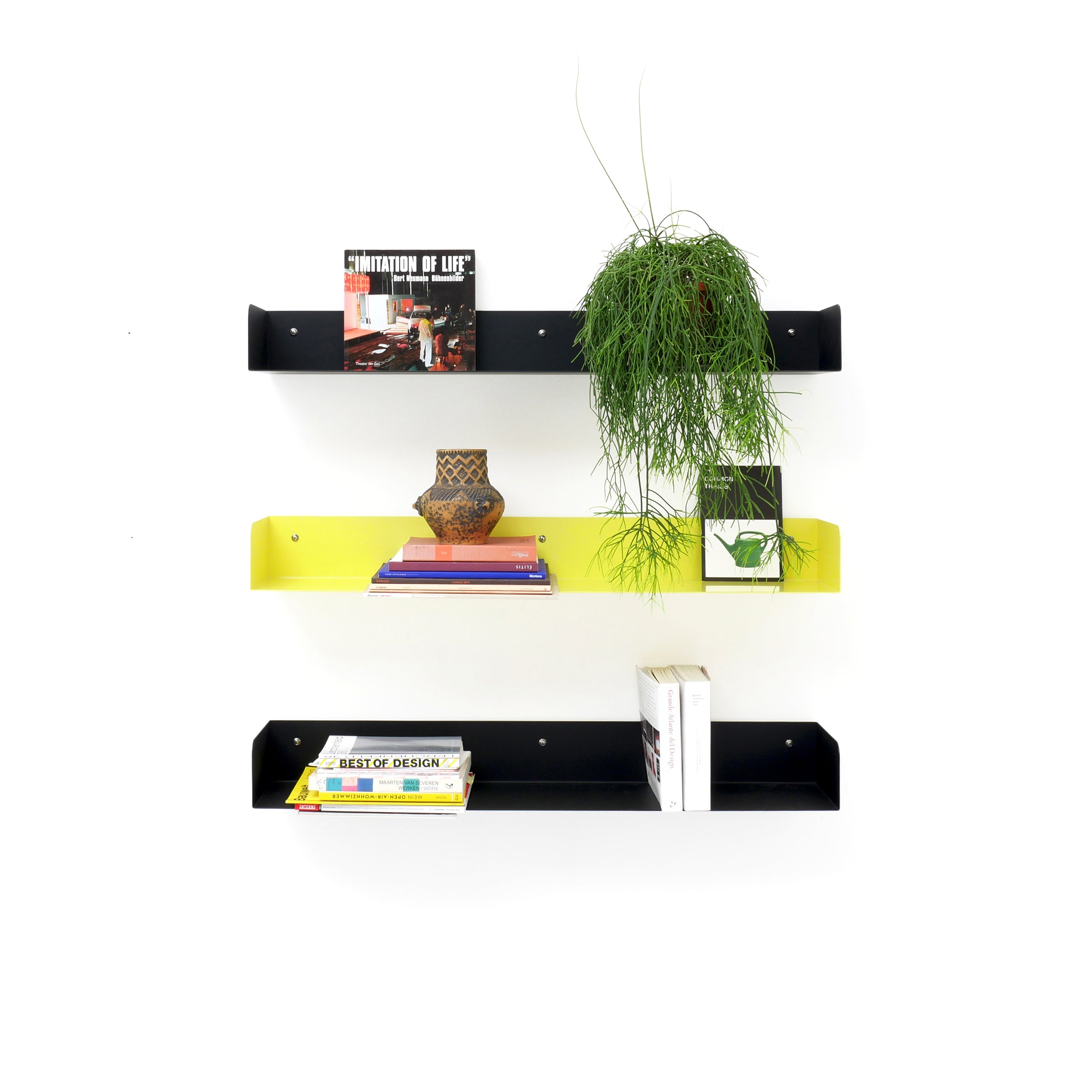 Three Atelier Haussmann Poggibonsi shelves: two in powder-coated black and one in vibrant yellow. The top shelf holds books and a plant, the middle has books, magazines, and a vase, while the bottom displays more books—a sleek addition against a crisp white background.